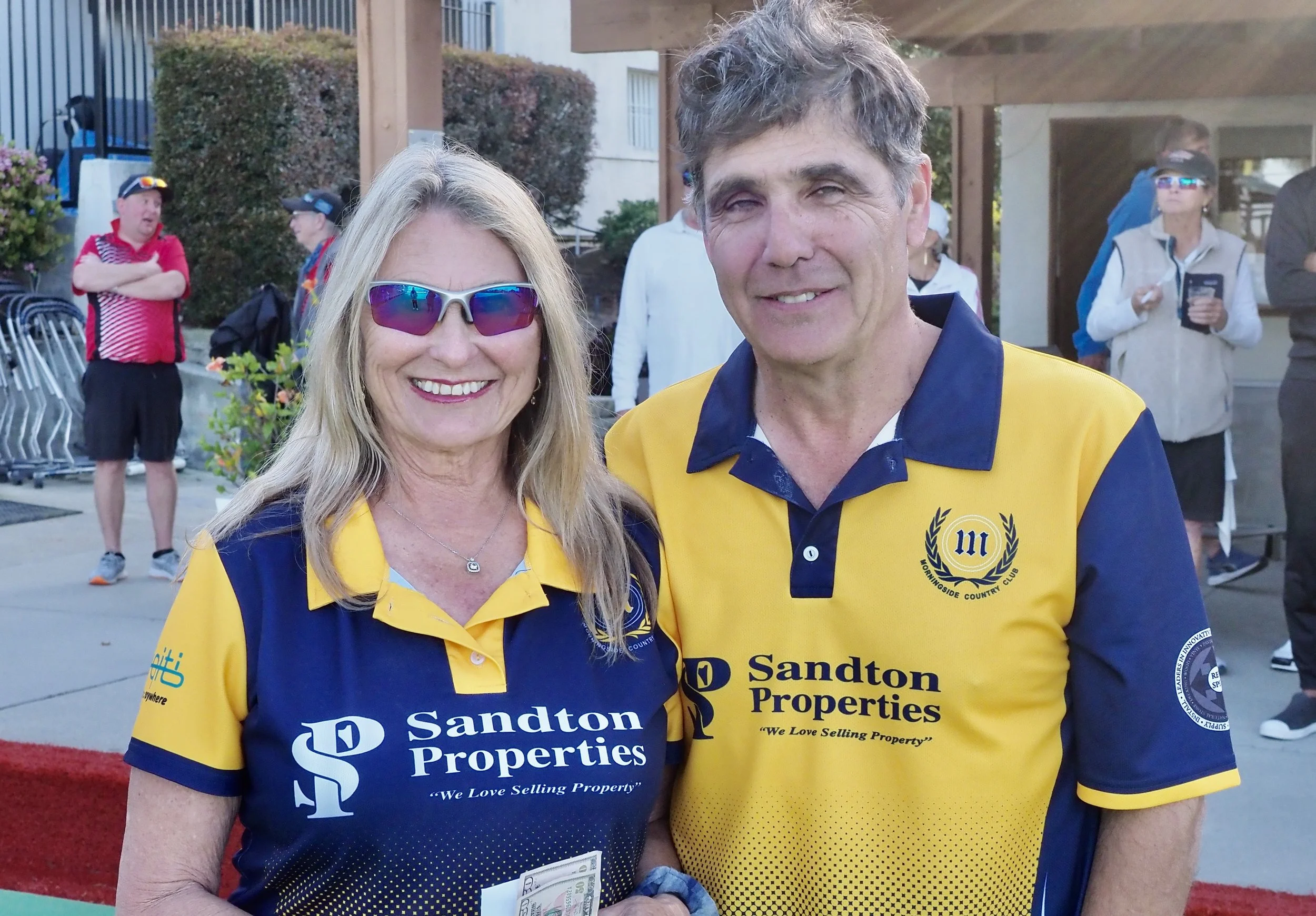 A smiling woman and man wearing matching yellow and blue Sandton Properties shirts, standing outdoors at a social or sporting event with other people in the background.