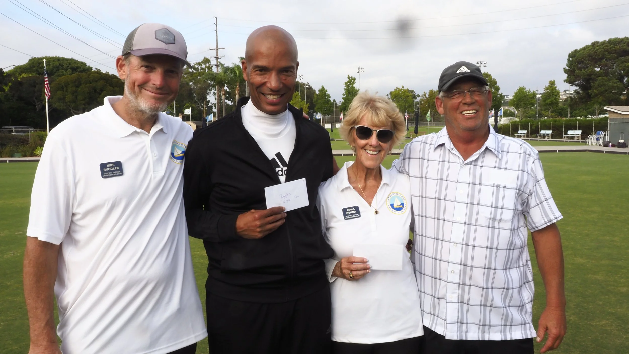 Four people standing outdoors on a lawn, smiling, holding envelopes, dressed casually and wearing badges and sunglasses. Background shows a fenced area and trees.