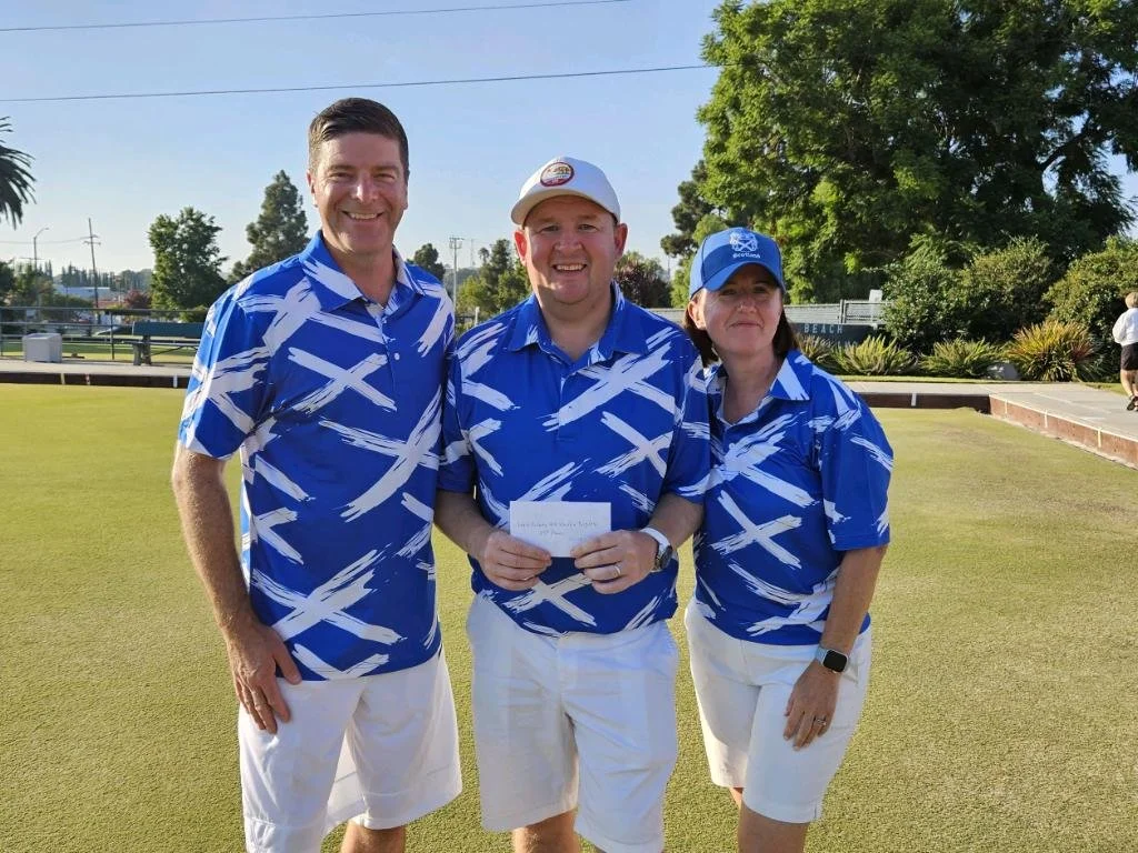 Three people in matching blue and white patterned shirts and white shorts standing on a lawn, smiling, with a tennis court and trees in the background.