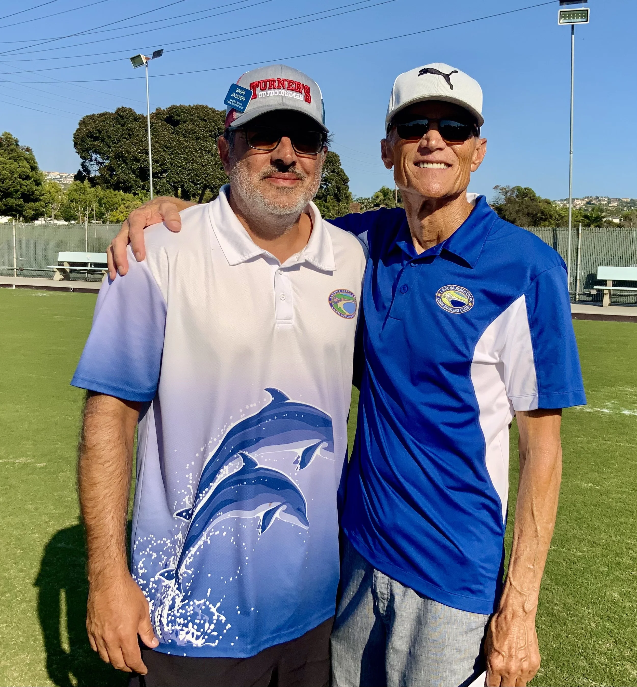 Two men standing on a grass field with a tennis court in the background, smiling and embracing each other. Both are wearing sports shirts with logos, sunglasses, and caps.