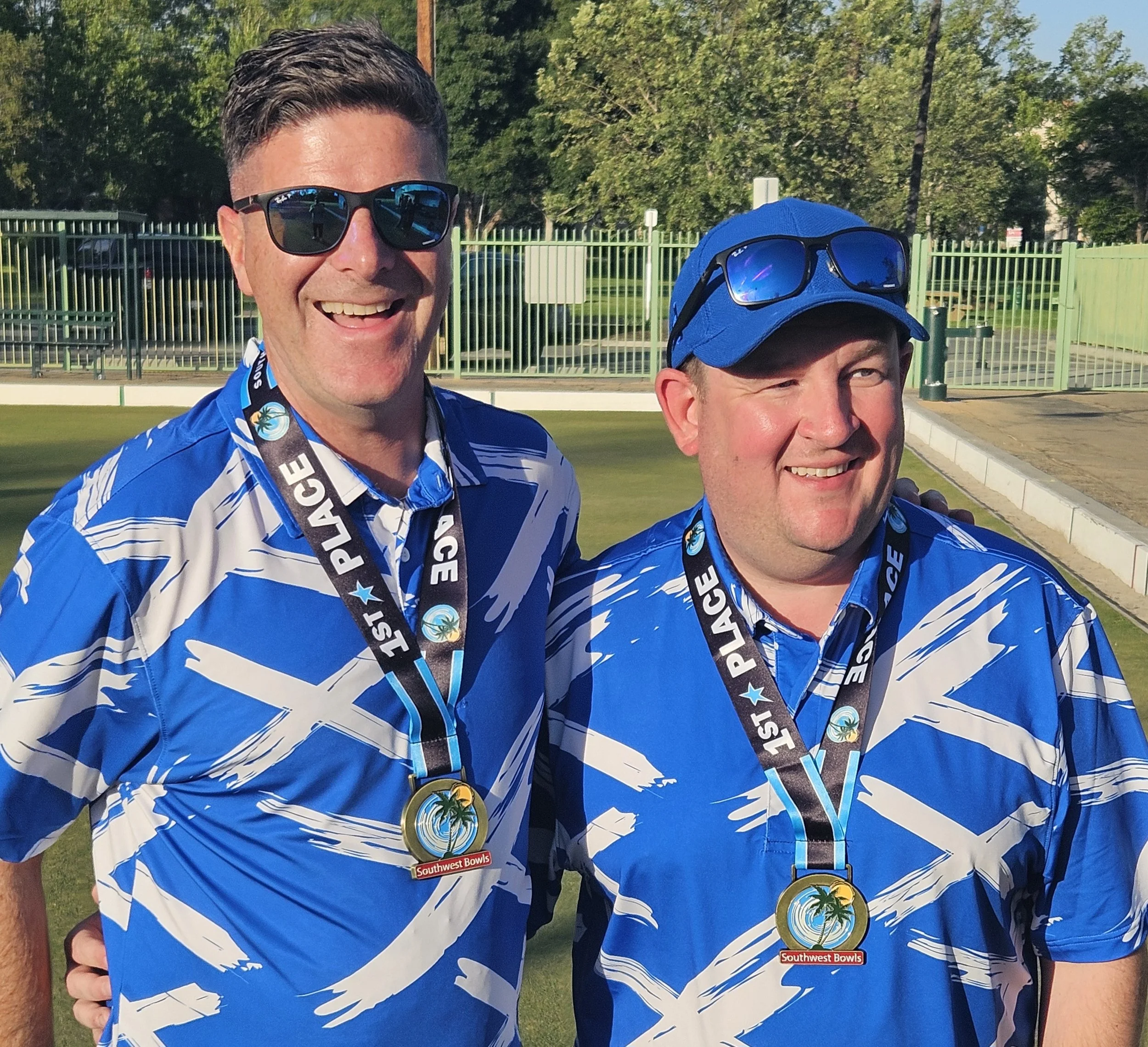 Two men wearing blue and white patterned shirts and medals around their necks, smiling outdoors on a sunny day, with a green fence and trees in the background.