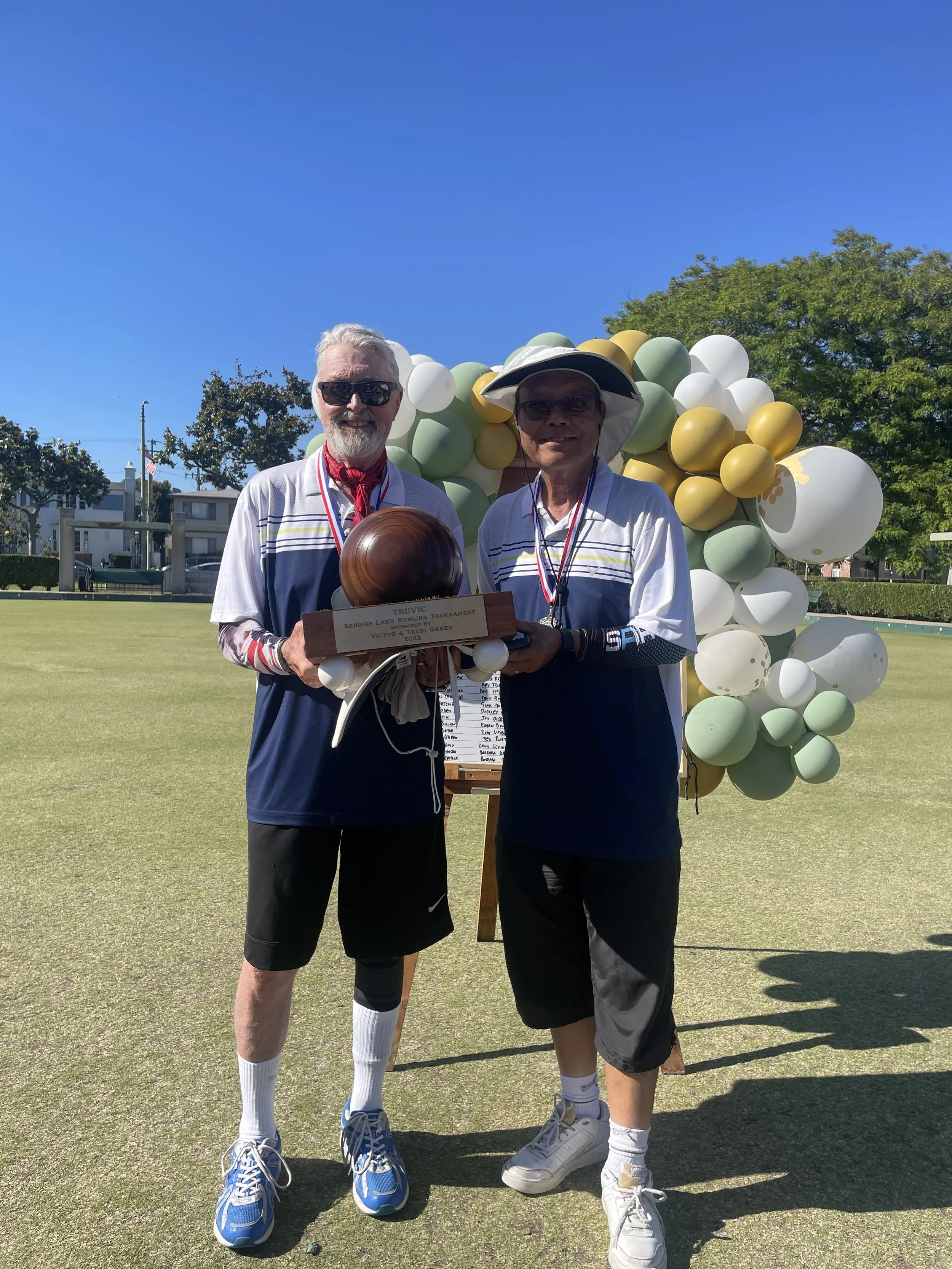 Two senior men standing on a lawn bowling green, holding a wooden trophy, wearing medals, and smiling. One man is wearing sunglasses and a white sun hat, the other has white hair and a red neckerchief. There is a large balloon decoration in the background and a blue sky overhead.