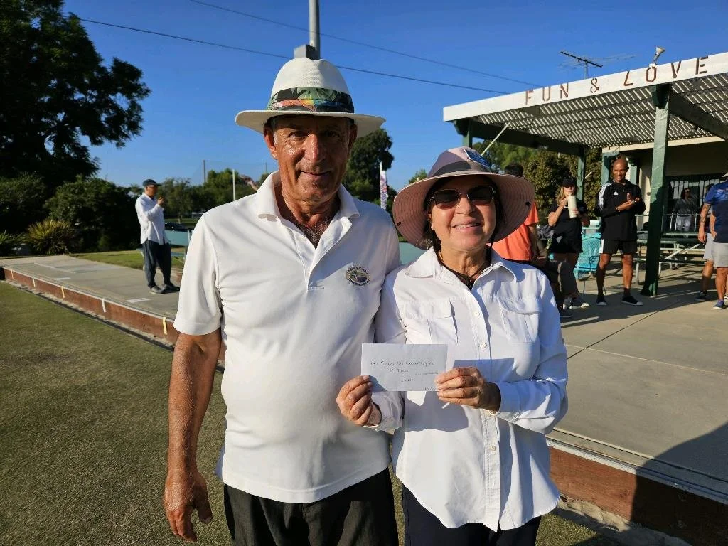 A man and woman wearing hats and white shirts standing outside, with the woman holding a white envelope, at a social gathering or event.