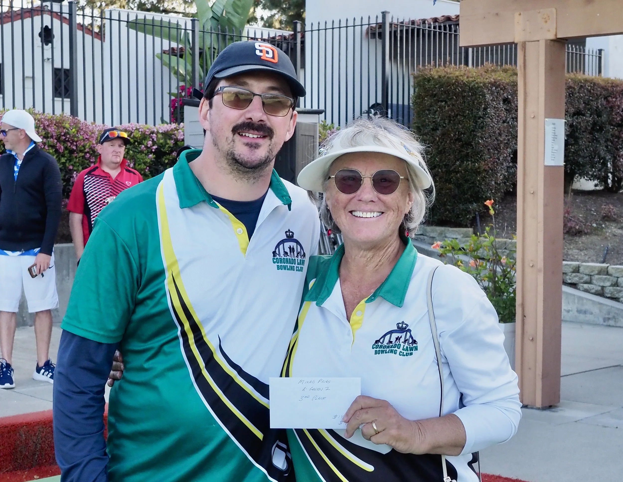 A man and woman, both wearing Coronado Lawn Bowling Club shirts, smiling outdoors at a lawn bowling event. The man wears glasses and a cap, the woman wears sunglasses and a hat. She is holding a note that says 'Mom Pavers, 2nd place'.