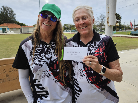 Two women standing outdoors, smiling at the camera. The woman on the right is holding a white paper. They are wearing matching black and white shirts with tiger designs, and the woman on the left is wearing sunglasses and a green cap.