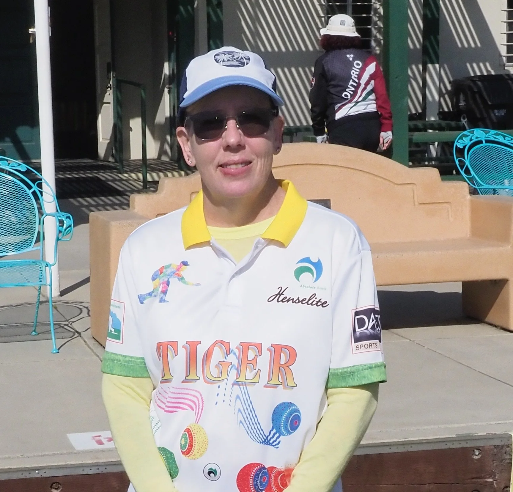 A woman wearing sunglasses, a white and yellow sports jersey with the word 'TIGER' and colorful graphics, and a white baseball cap, sitting outdoors on a bench.