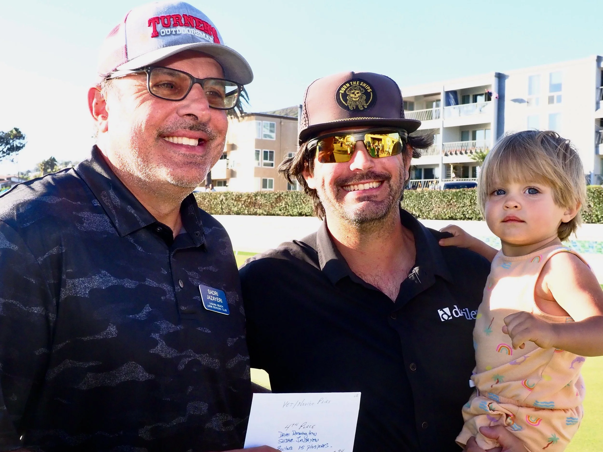 Three people smiling outdoors, two men and a young girl, with buildings in the background.