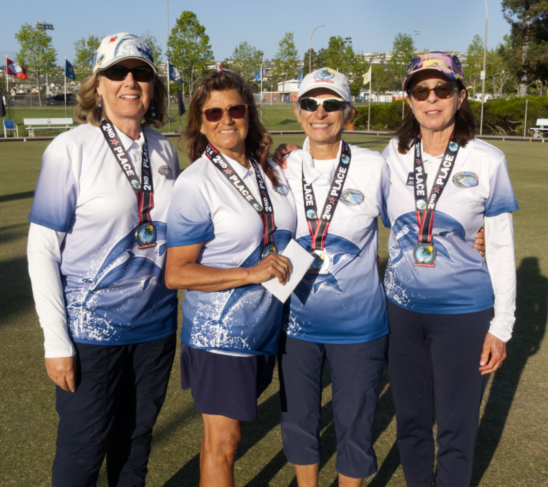 Four senior women standing on a lawn, wearing matching blue and white shirts with a water and wave design, and medals around their necks that say '2nd Place', smiling at the camera.