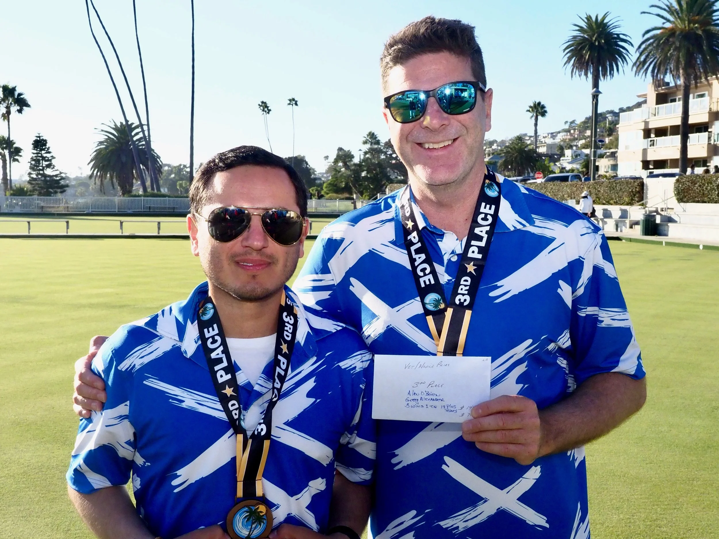 Two men wearing blue and white patterned shirts and sunglasses standing on a lawn with palm trees, buildings, and a clear sky in the background, celebrating their third place win in a competition.