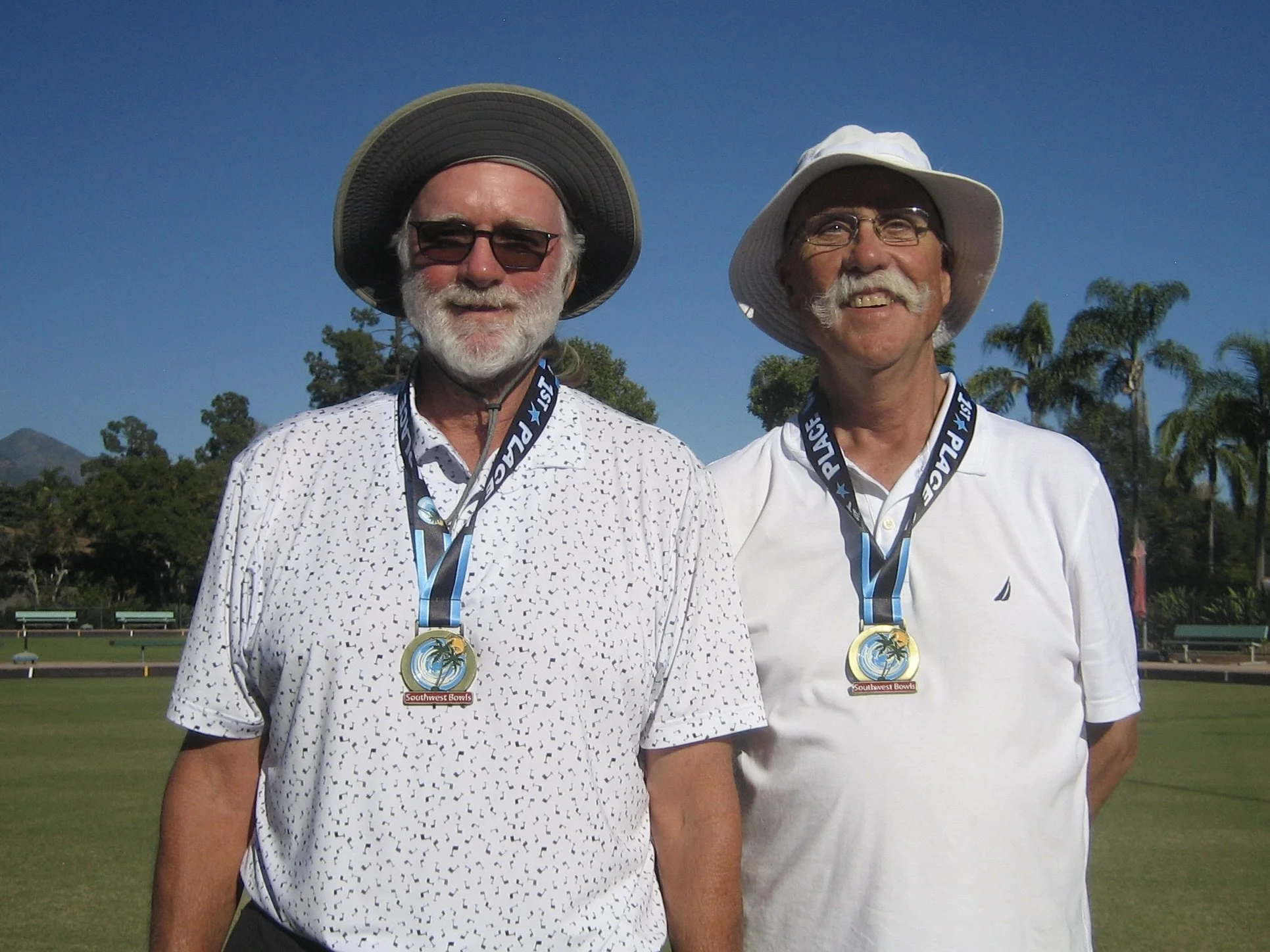 Two smiling older men wearing white shirts and sun hats, standing outdoors on a grassy field with tennis courts and palm trees in the background, each with a medal around their neck reading 'Southwest Bowls'.