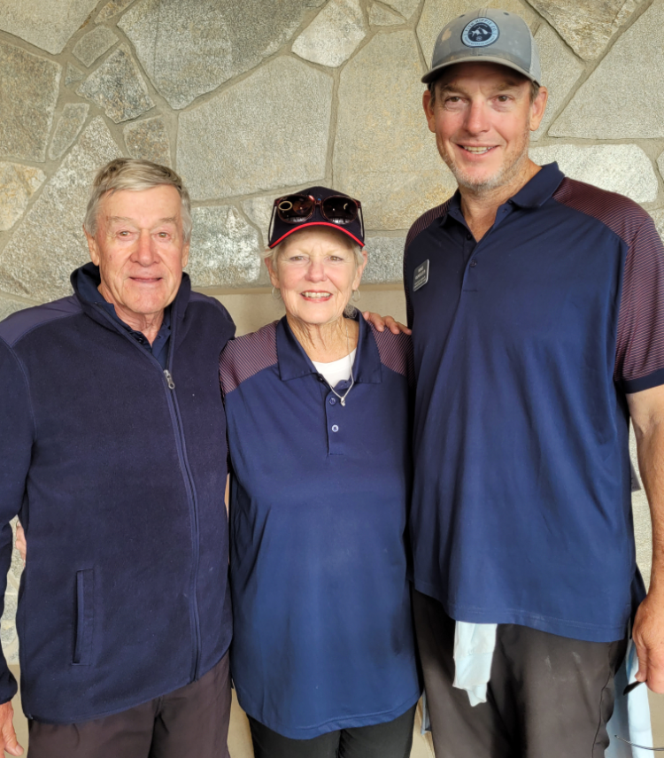 Three smiling adults, two men and one woman, standing together indoors against a stone wall. The woman is in the middle, wearing a navy blue polo shirt and a cap with sunglasses on top. The man on the left is wearing a dark jacket, and the man on the right is tall, wearing a blue polo shirt and a gray cap.