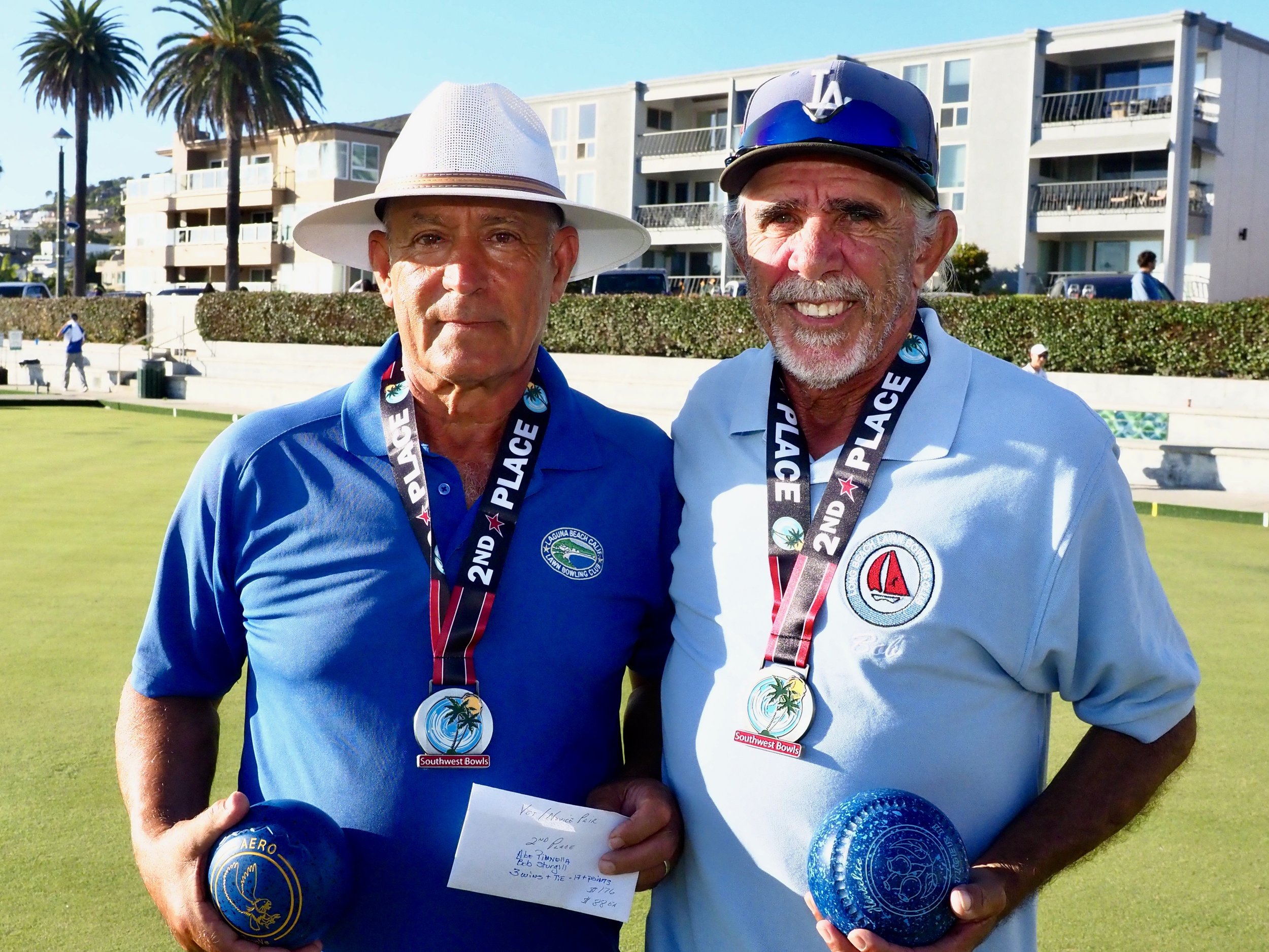 Two men standing on a lawn holding medals, one wearing a white hat and a blue shirt, the other wearing a helmet and a light blue shirt, both smiling, with houses and palm trees in the background.