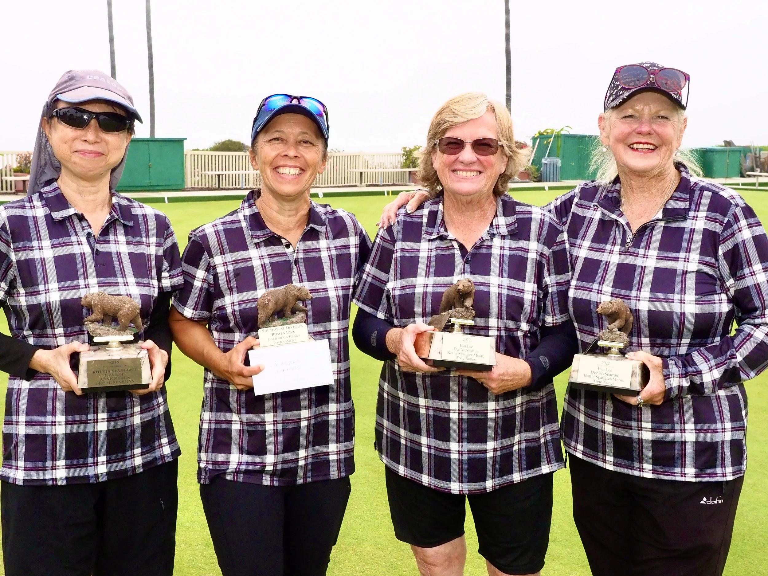 Four women standing on a grassy field, smiling, holding trophies of bears on bases, wearing matching purple plaid shirts, sunglasses, and hats.