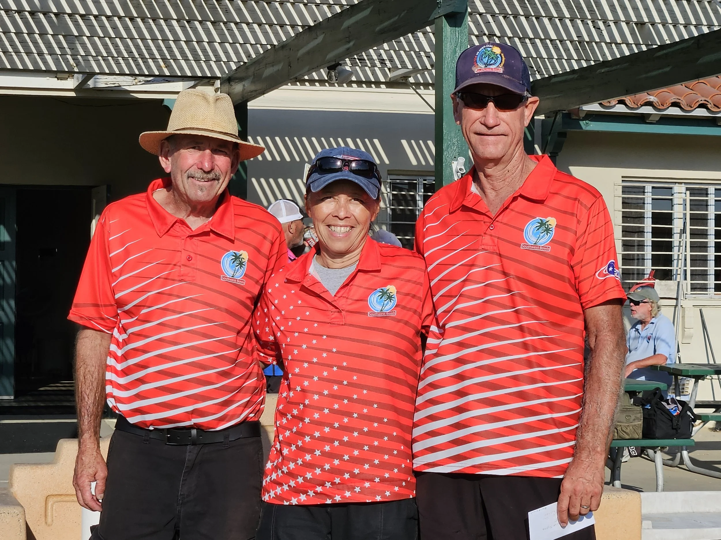 Three smiling people wearing matching red striped shirts with tropical logos at a bowling event, standing outdoors with a building and a woman in the background.
