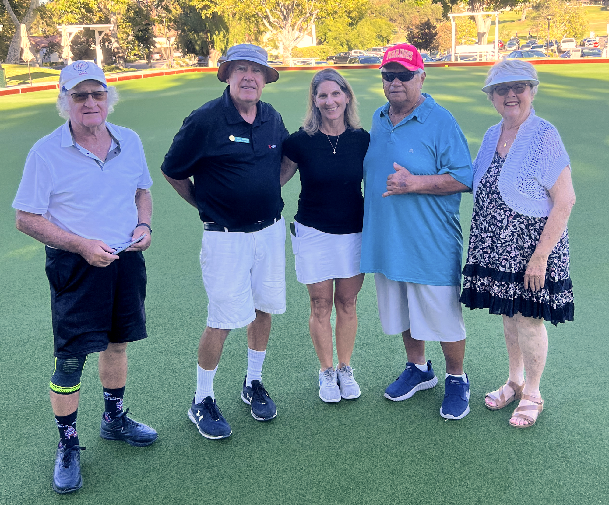 Five older adults standing on a mini golf course, smiling at the camera. The background shows trees, parked cars, and parts of the course with barriers. They are dressed casually, some wearing hats and sunglasses.