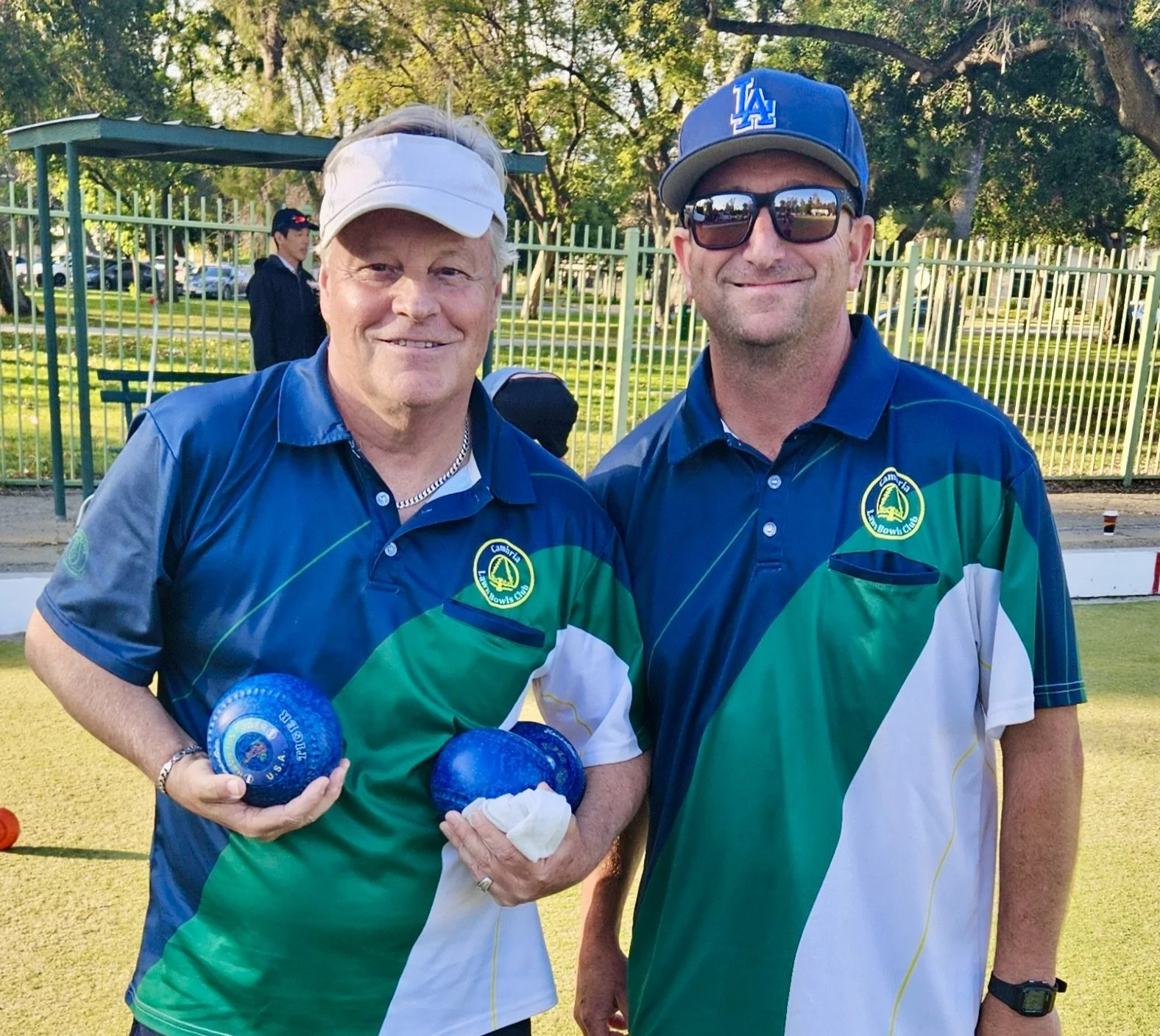 Two men wearing matching green and blue shirts, standing outdoors on a lawns bowling green, smiling at the camera. One holds several blue lawn bowls.