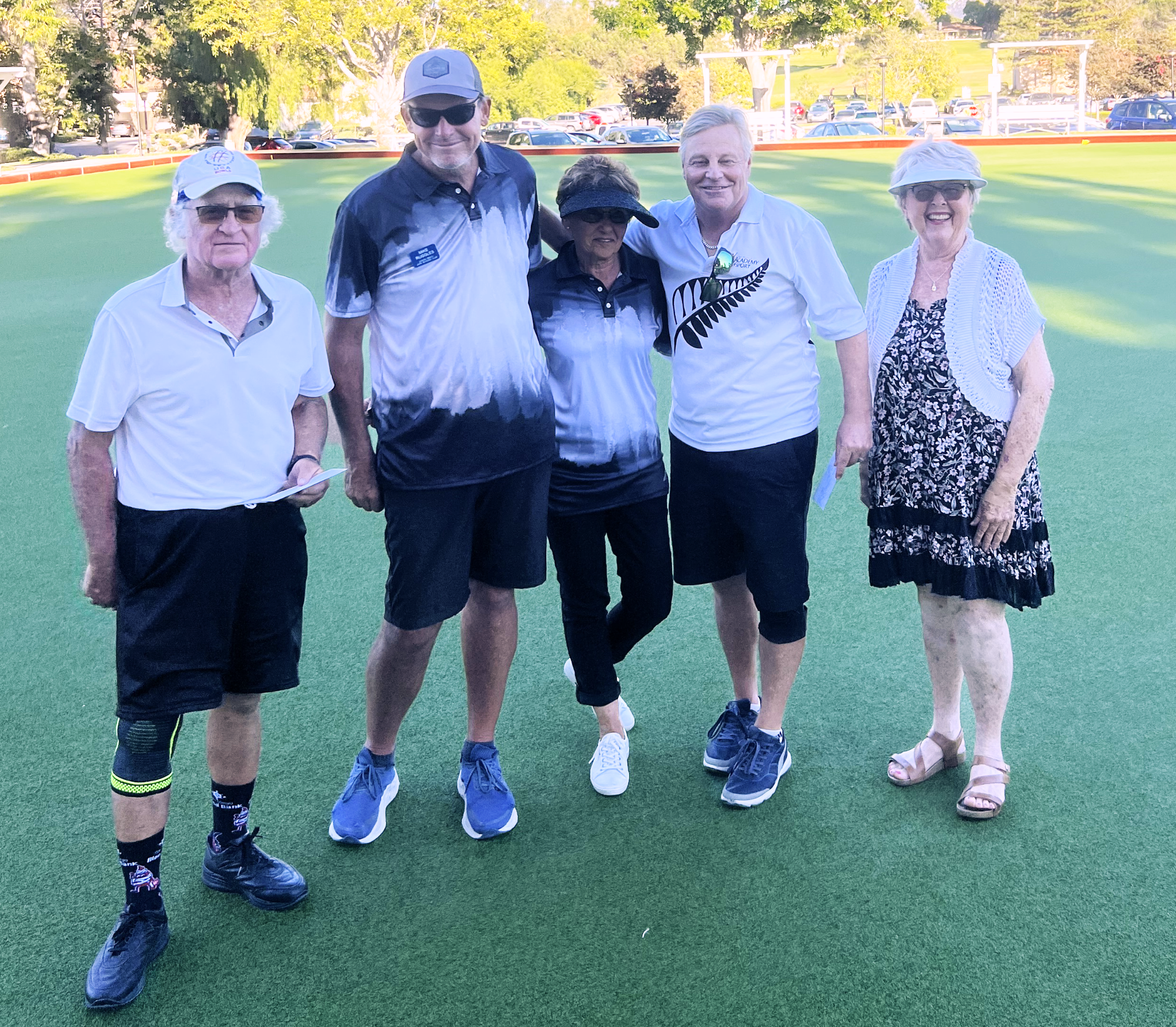 Five older adults standing on a green sports field, smiling, dressed in casual summer clothes, with trees and parked cars in the background.