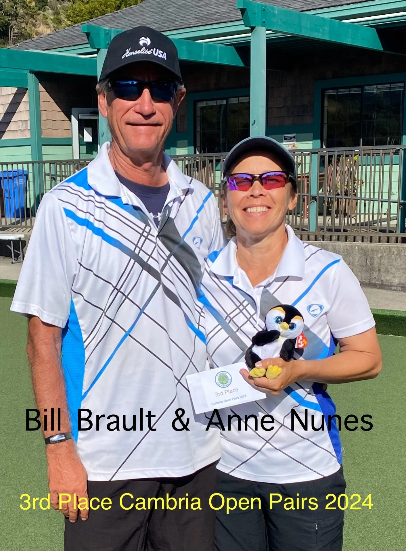 Bill Brault and Anne Nunes in matching tennis outfits at the Cambria Open Pairs 2024, holding a third-place award and a plush penguin mascot, standing on a tennis court with a building in the background.