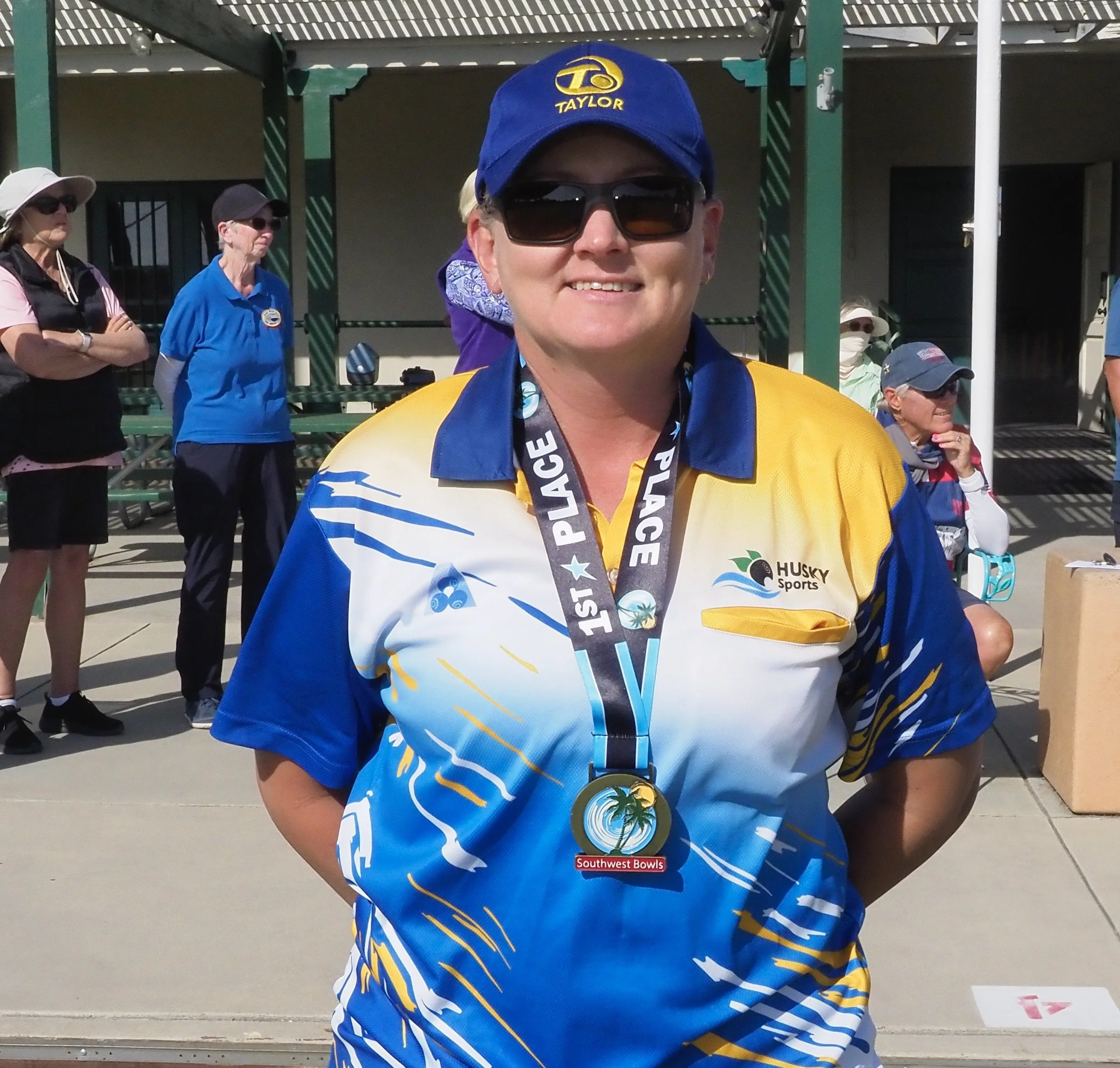 A woman wearing sunglasses, a blue and yellow sports jersey, and a blue cap stands outdoors with a gold medal around her neck that reads 'SWBC Southwest Bowls.' She has a smile on her face, and there are other people in the background, some wearing hats and casual sports attire.