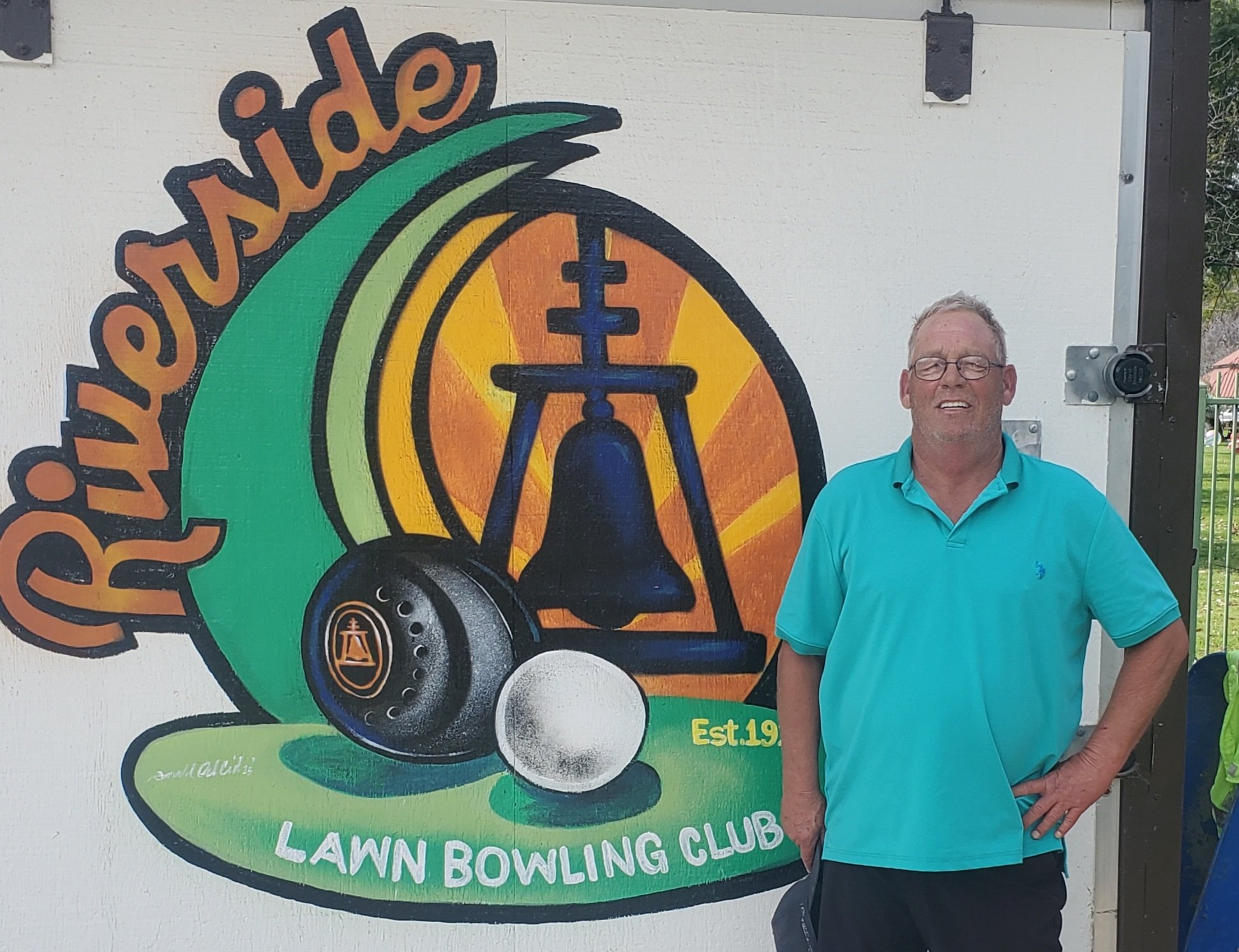 Man wearing glasses and a turquoise polo shirt standing next to a colorful sign for the Riverside Lawn Bowling Club. The sign features a large bell, a lawn bowling ball, and a small logo with a bell in front of a sunset background, with the text "Riverside Lawn Bowling Club" and "Est. 19".