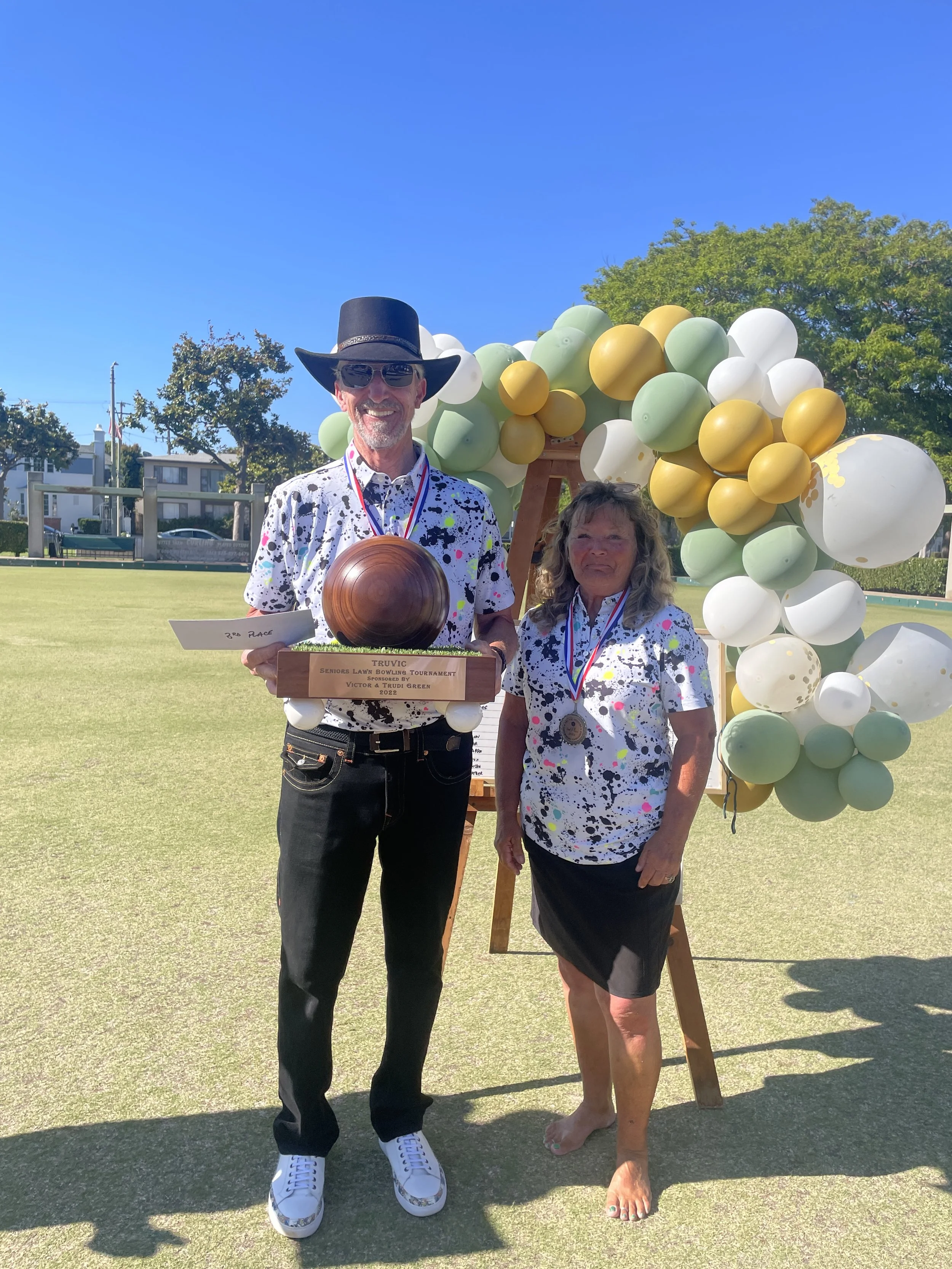 Two people standing on a lawn holding trophies, wearing medals and matching paint-splattered shirts, with a large balloon arch behind them. The man is wearing a black wide-brimmed hat and sunglasses, and the woman is barefoot.