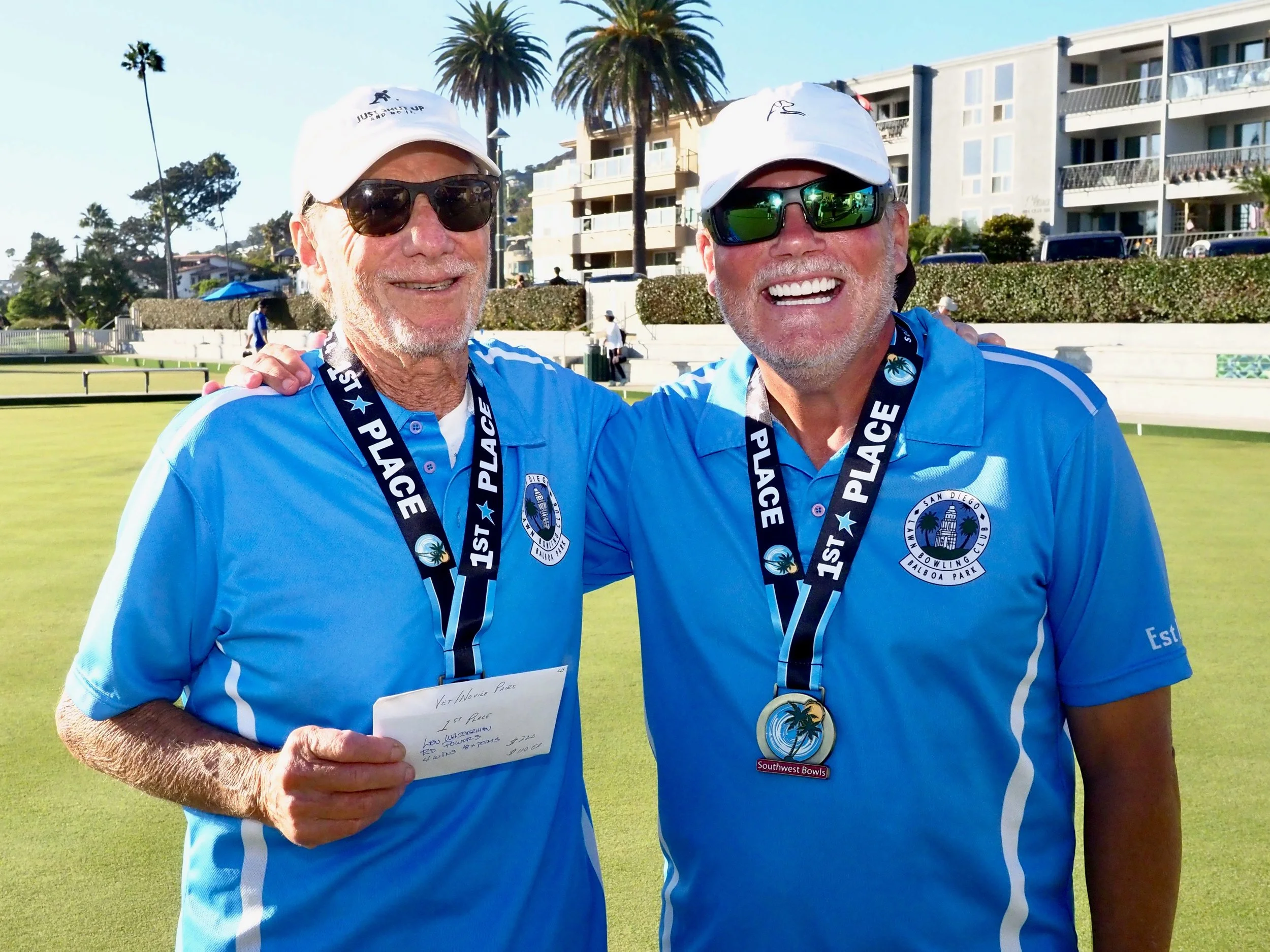 Two men wearing blue polo shirts and medals with '1st Place' ribbons around their necks, smiling on a lawn bowling green with palm trees and apartment buildings in the background.
