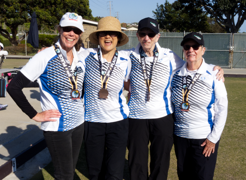 Four women standing outdoors, wearing matching sports shirts, black pants, and medals around their necks, smiling for a photo during a sporting event.