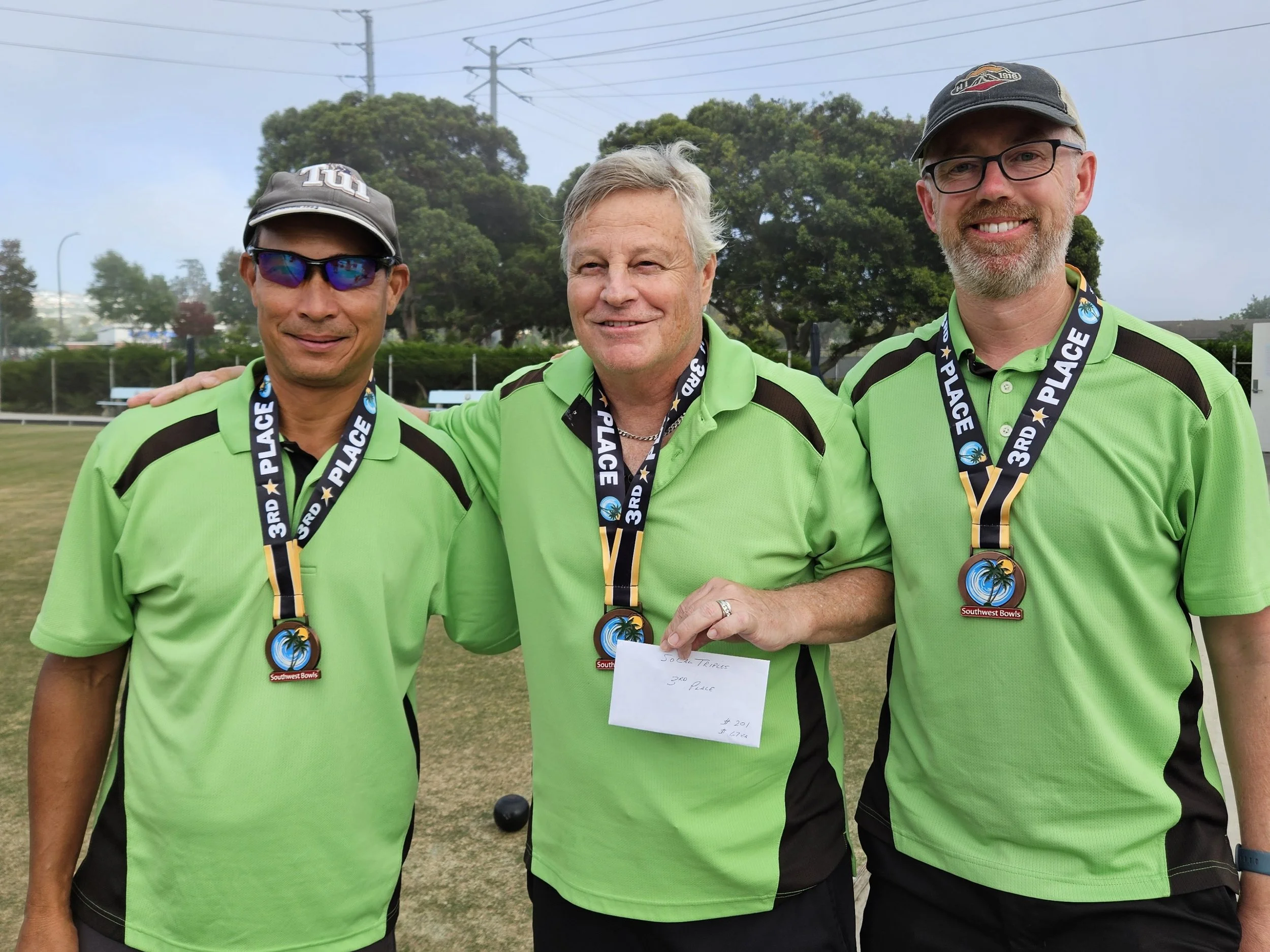 Three men in green sports shirts with medals around their necks, standing outdoors on a field, smiling and posing for a photo.