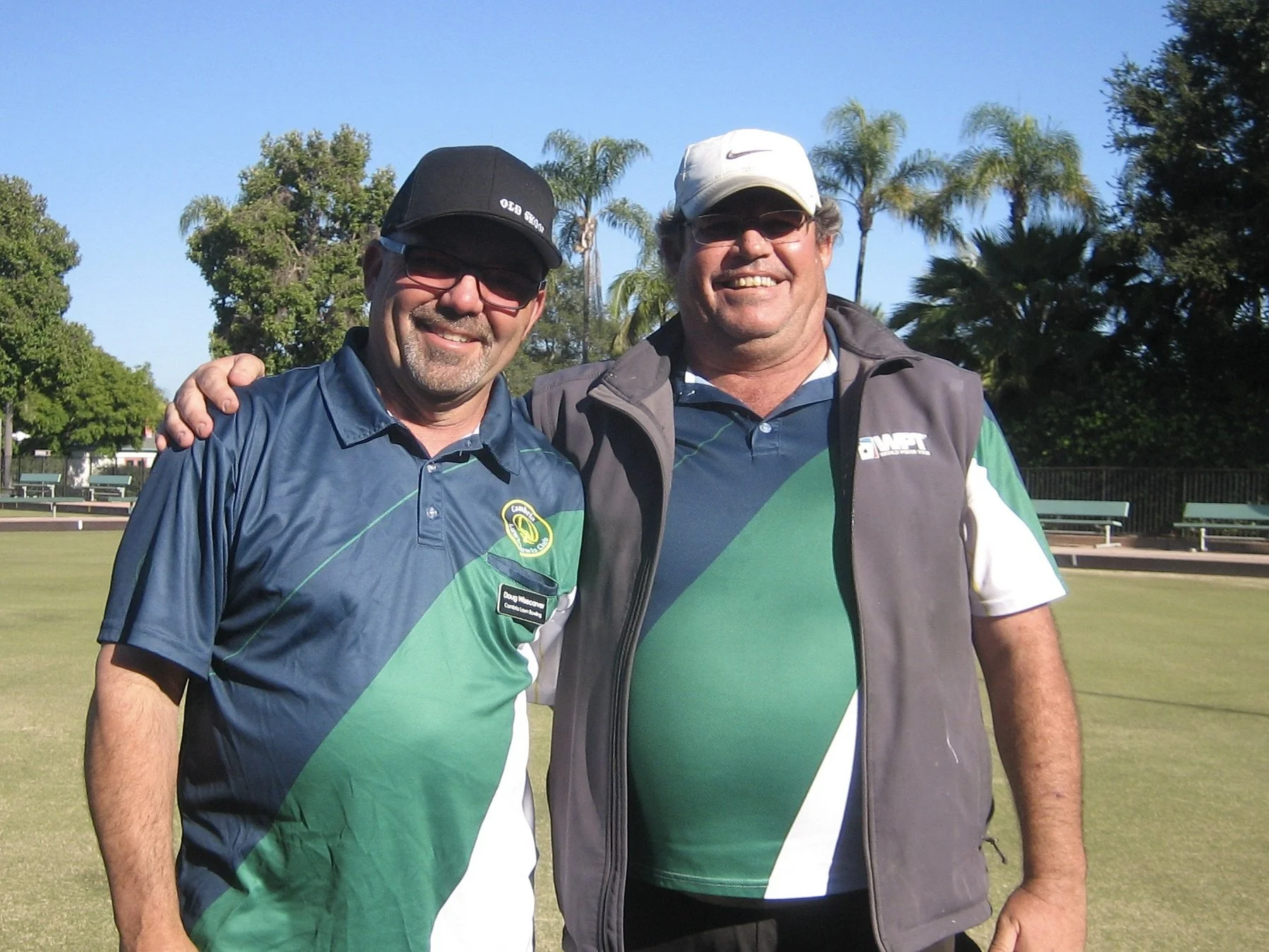 Two middle-aged men smiling and standing outdoors on a golf course with trees and clear blue sky in the background. One man is wearing a black cap and glasses, while the other is wearing a white cap.