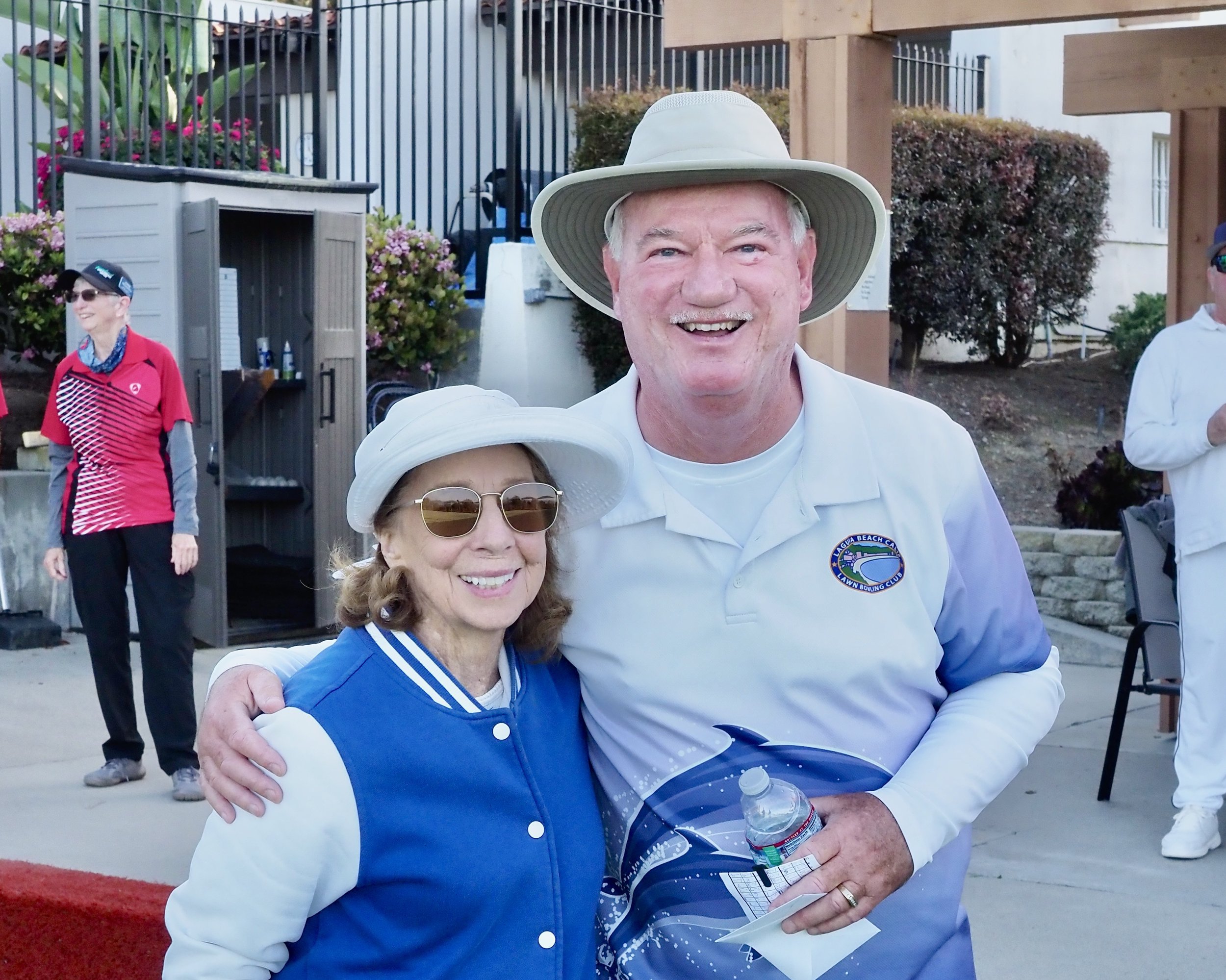 An elderly man wearing a wide-brimmed hat and white lapel shirt with a logo, and an elderly woman in sunglasses and a white hat, hugging and smiling at an outdoor event.