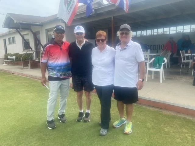 Four people standing on a golf course, smiling at the camera, with buildings and flags in the background.
