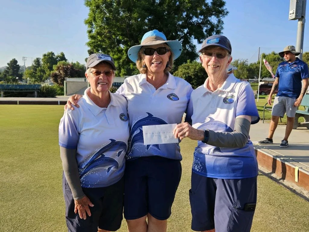 Three women in matching white and blue shirts posing for a photo on a bocce court, with one woman in the middle holding a check. A man is in the background near the court.