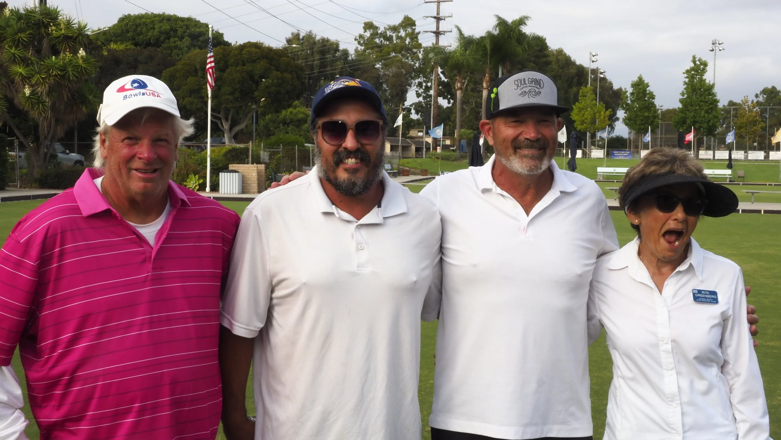 Four people standing together outdoors, smiling, on a grassy field with trees, fences, and flags in the background. They are dressed in casual clothing, including hats and sunglasses.