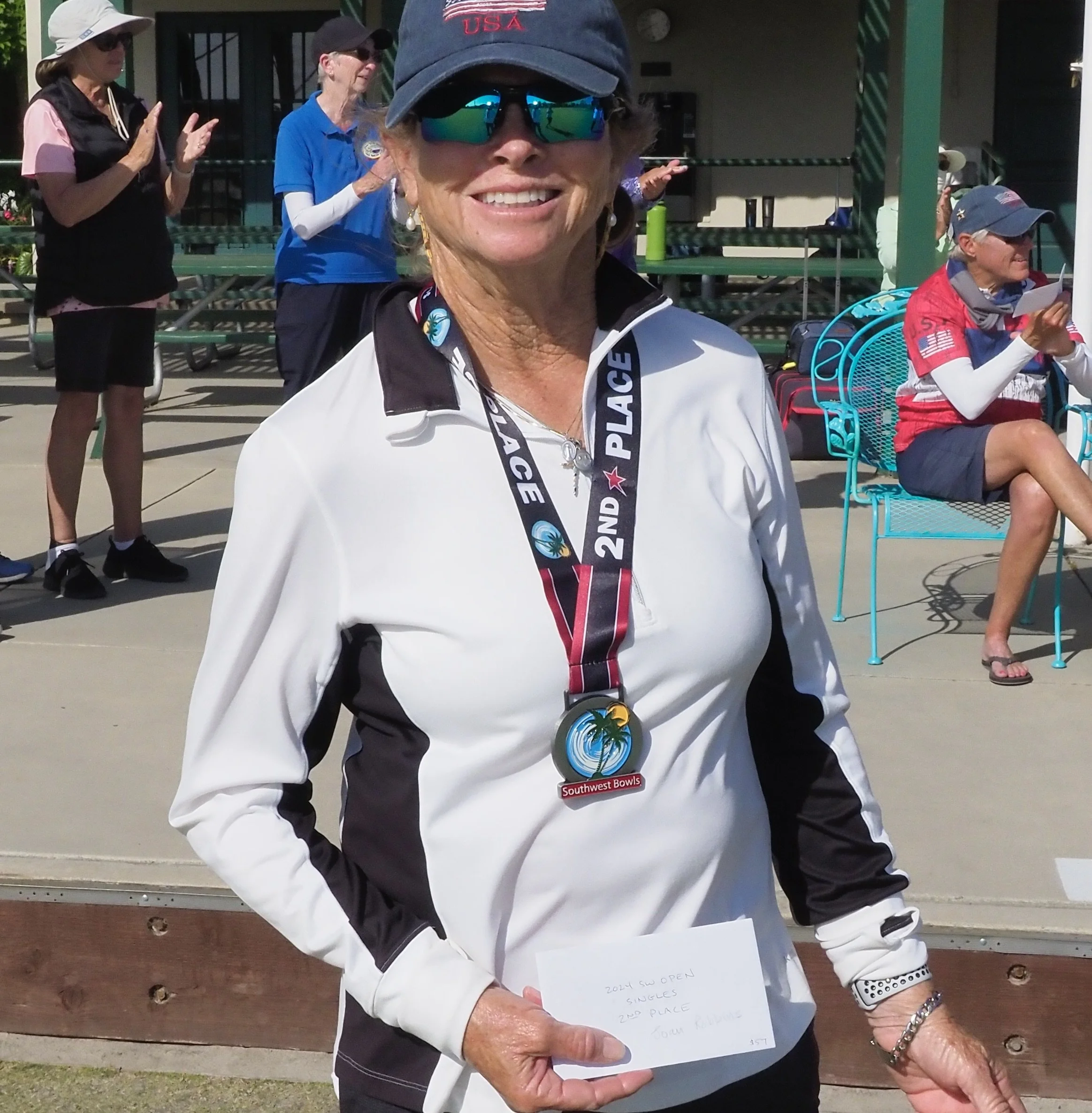 A woman wearing sunglasses, a cap with 'USA' embroidered on it, and a white and black sports jacket, holding a medal and a piece of paper, standing outdoors at a sports event with other people in the background.