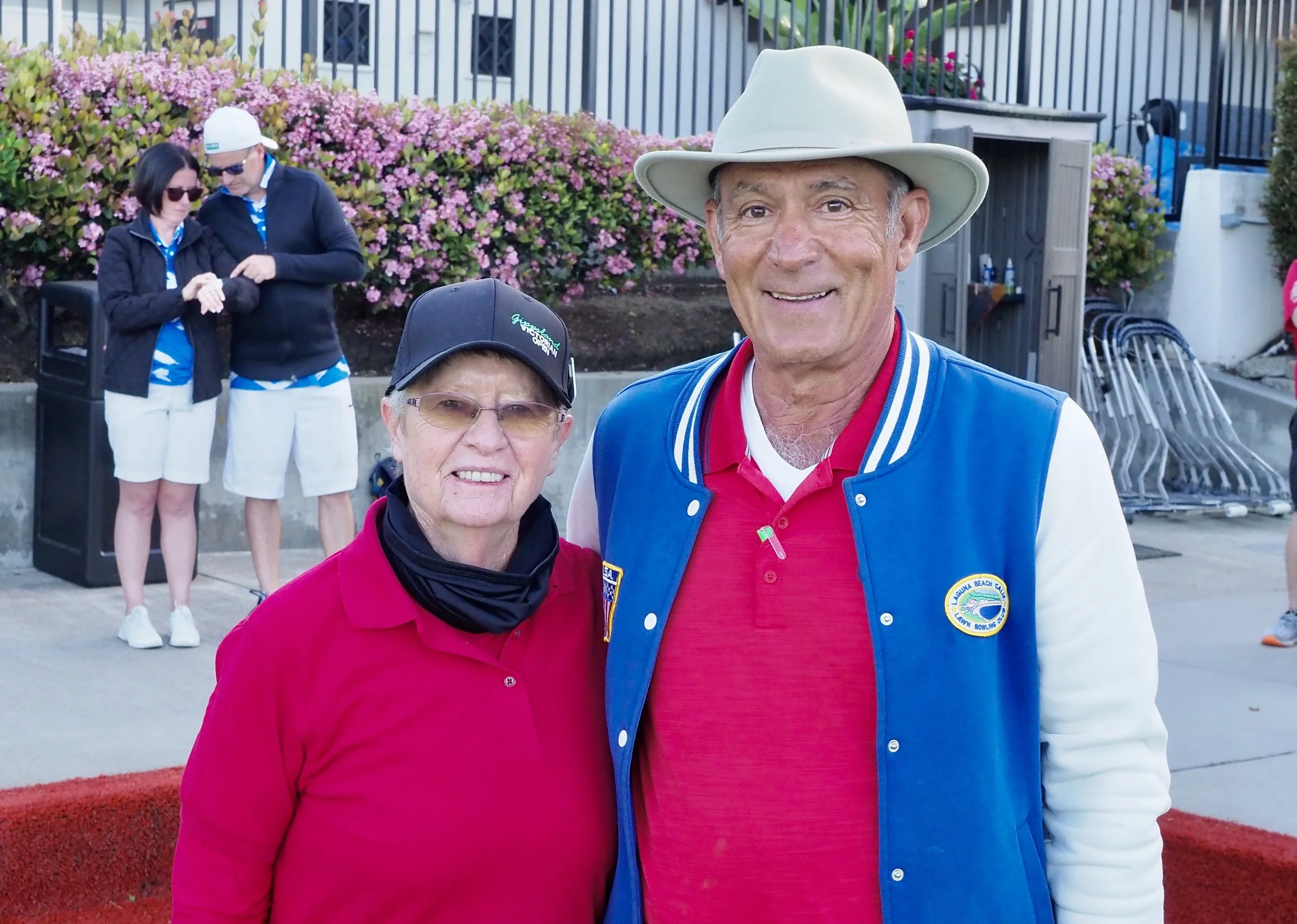 Two older individuals posing outdoors, the woman in a red shirt and black cap, the man in a red polo and a blue letterman jacket with a white hat, at a lawn bowling event with pink flowers and other people in the background.