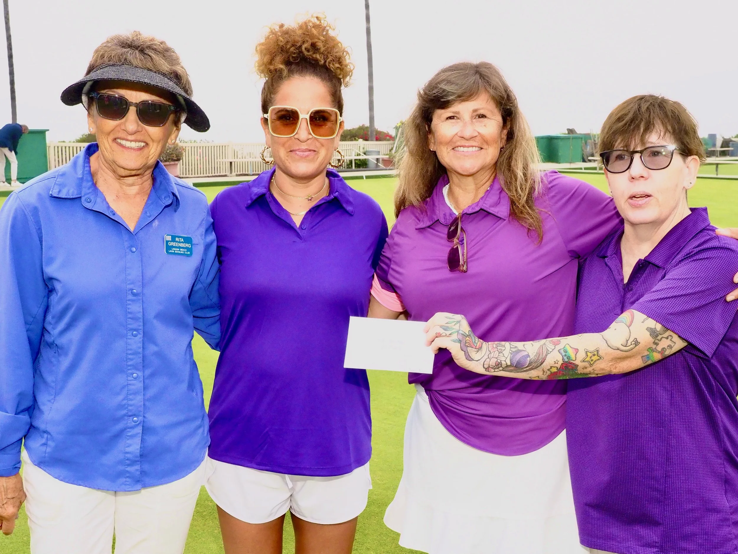 Four women in purple and blue shirts standing outdoors on a grassy field, one holding a white check.