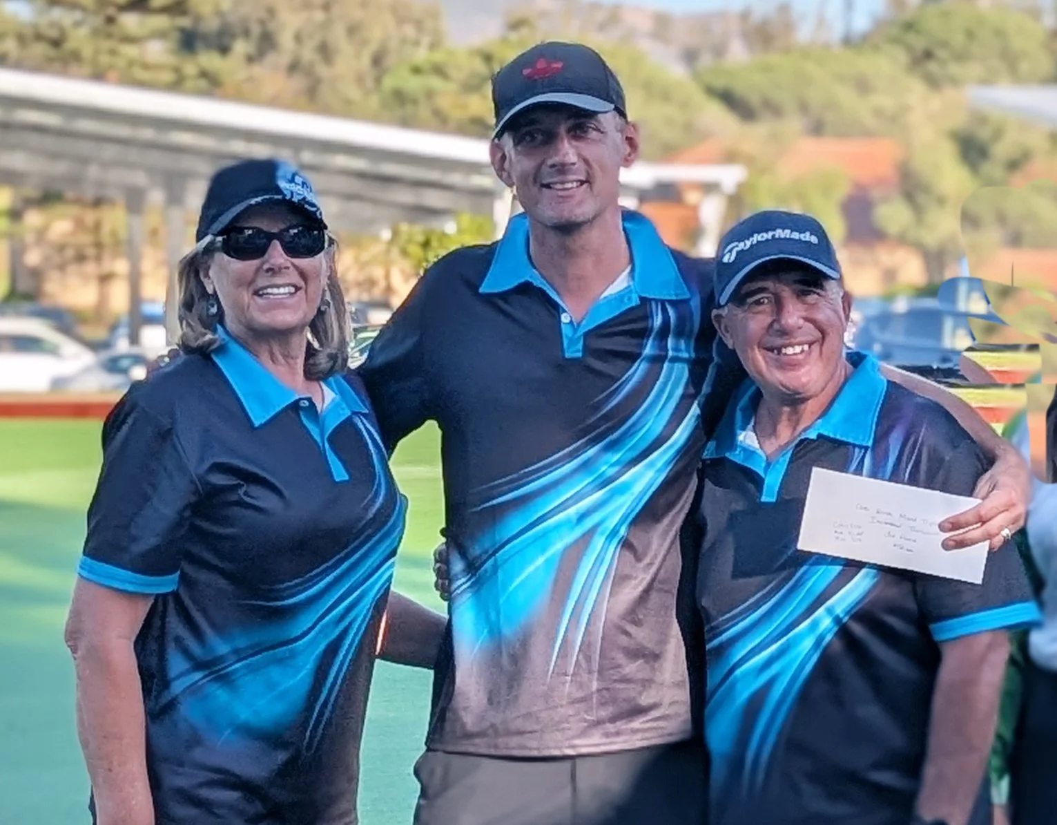 Three people in golf attire smiling and posing outdoors on a golf course, two women and one man, holding an envelope.