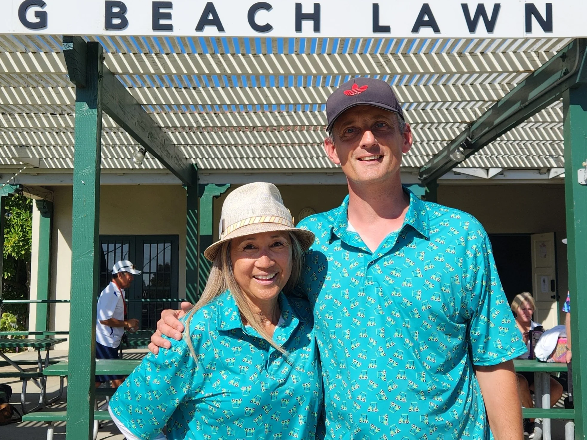 A smiling woman and man in matching blue patterned shirts standing under a sign that says G BEACH LAWN, with other people and picnic tables in the background.