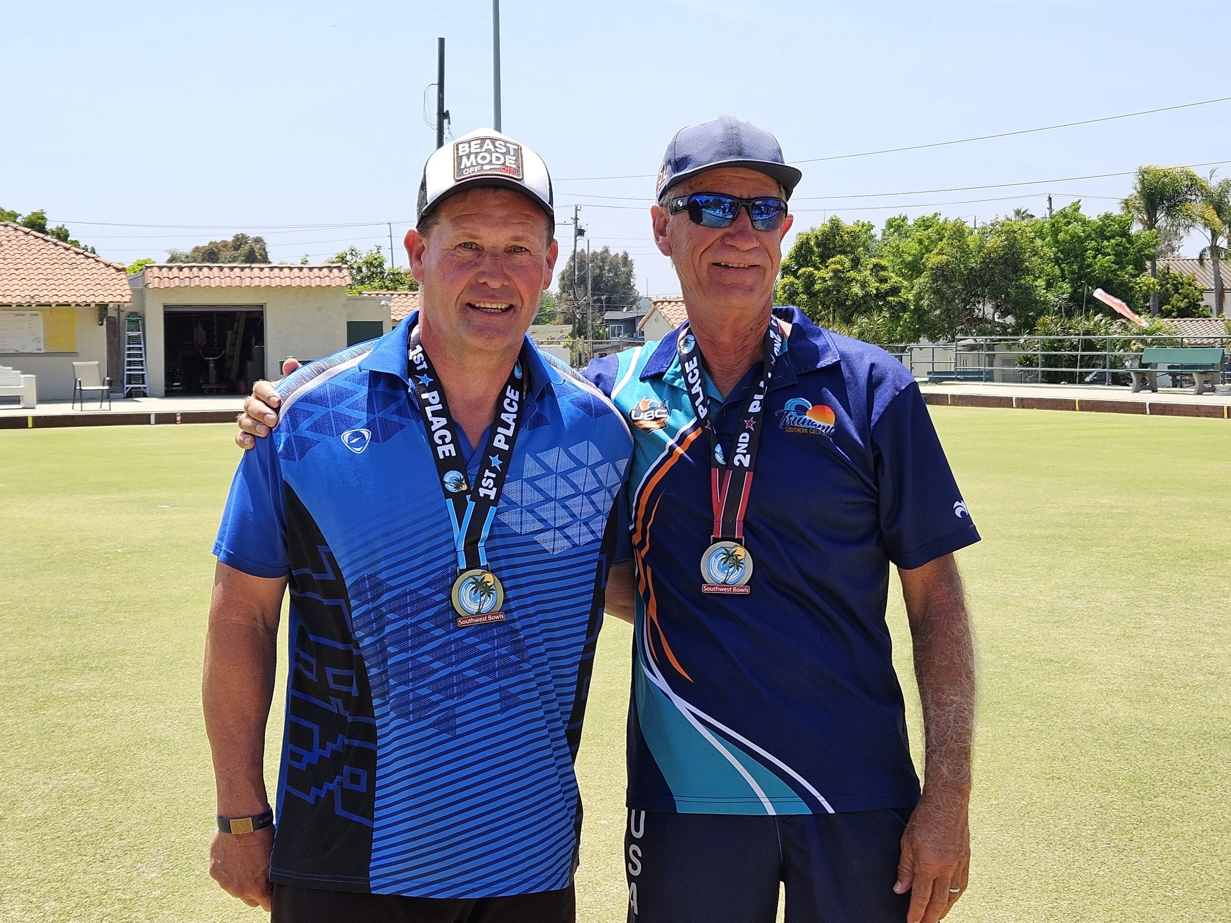 Two men standing on a green lawn, wearing medals and sports shirts, smiling after a competition, with trees, buildings, and a clear sky in the background.