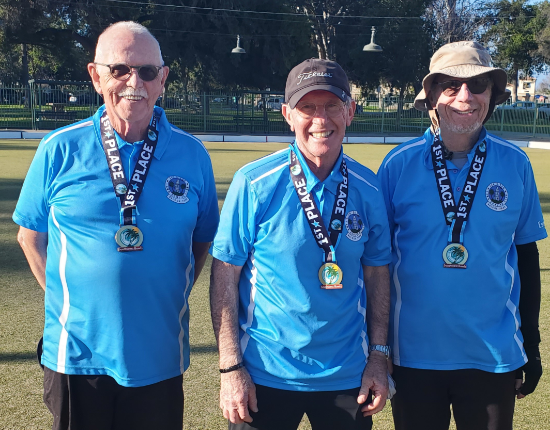 Three senior men standing outdoors on grass, wearing blue sports jerseys and medals around their necks, smiling at the camera.