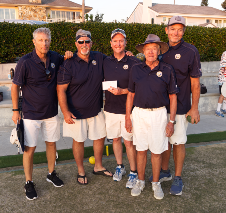 Five men in matching navy blue shirts and white shorts standing outdoors on a sunny day at a golf course, posing for a group photo with happy expressions.