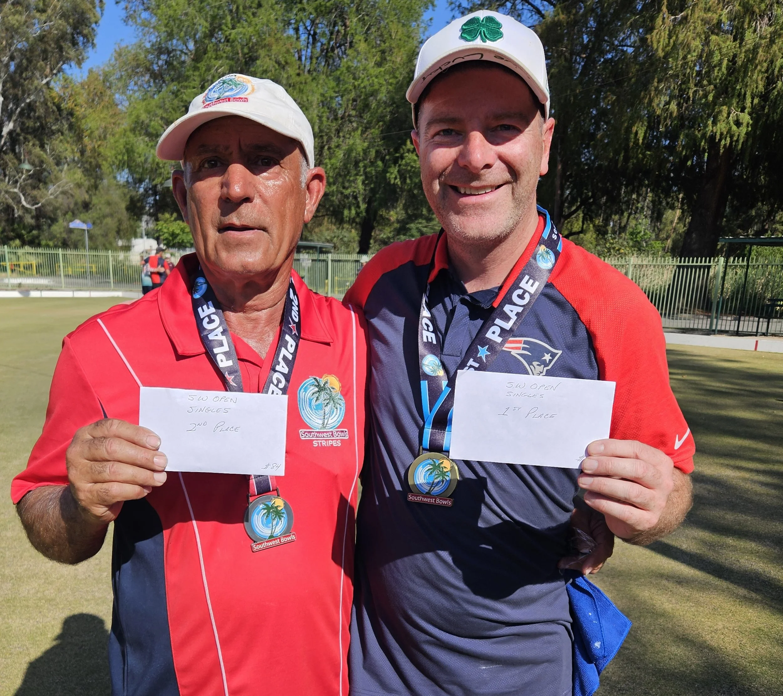 Two men in red and navy shirts, wearing medals, smiling on a golf course during daytime. They hold handwritten notes indicating their placements in a golf tournament, with trees and a fence in the background.