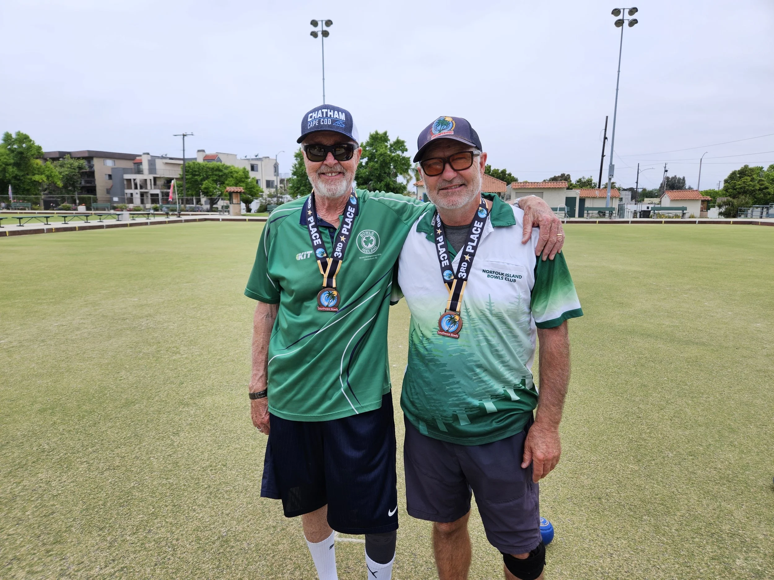 Two men standing on the bowls green with medals around their necks, smiling, arm around each other's shoulders, wearing green and white sports shirts, caps, and sunglasses.