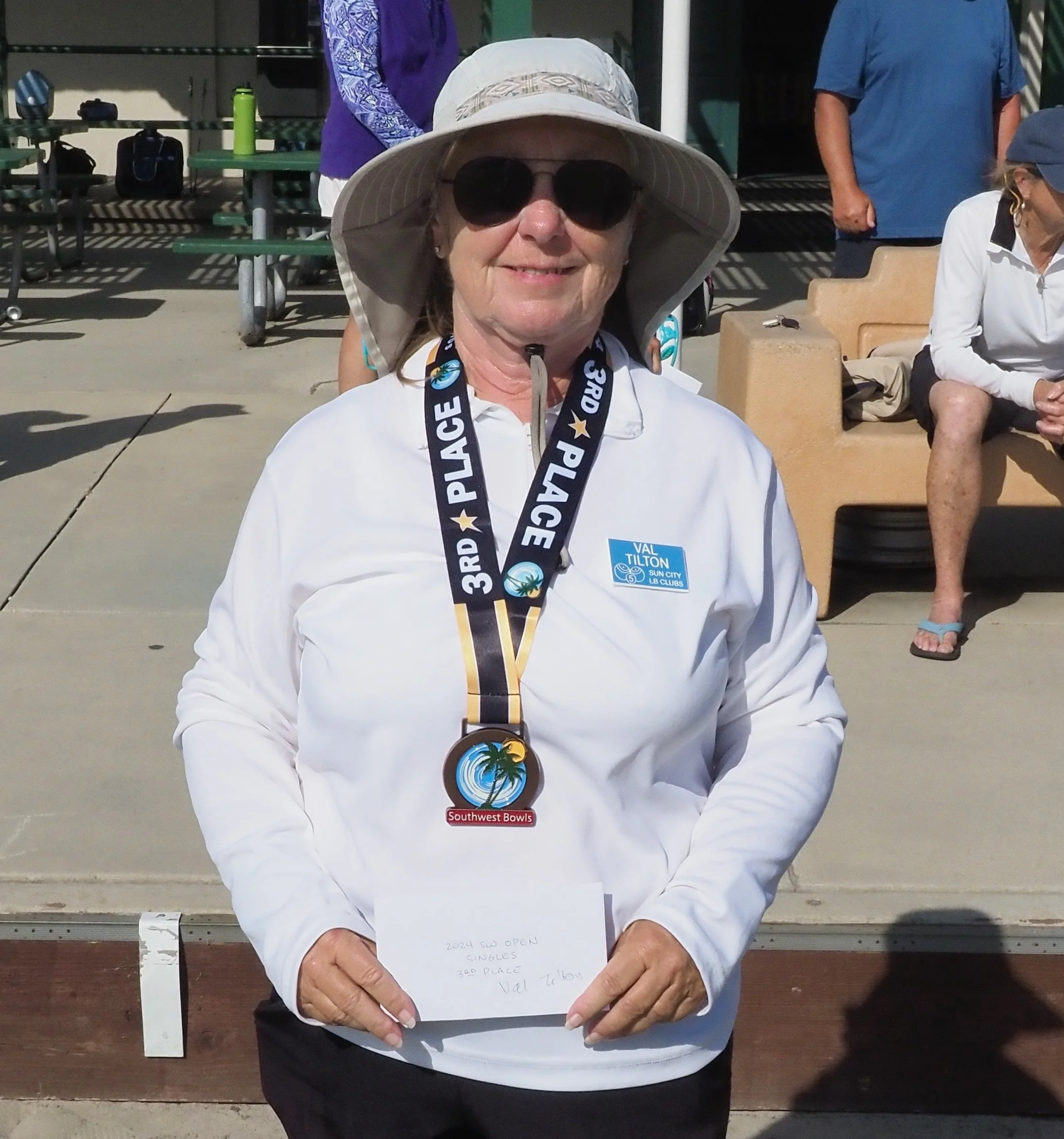 Woman wearing sunglasses and a large sun hat, holding a note and medal for third place at an outdoor event, with other people sitting and standing in the background.