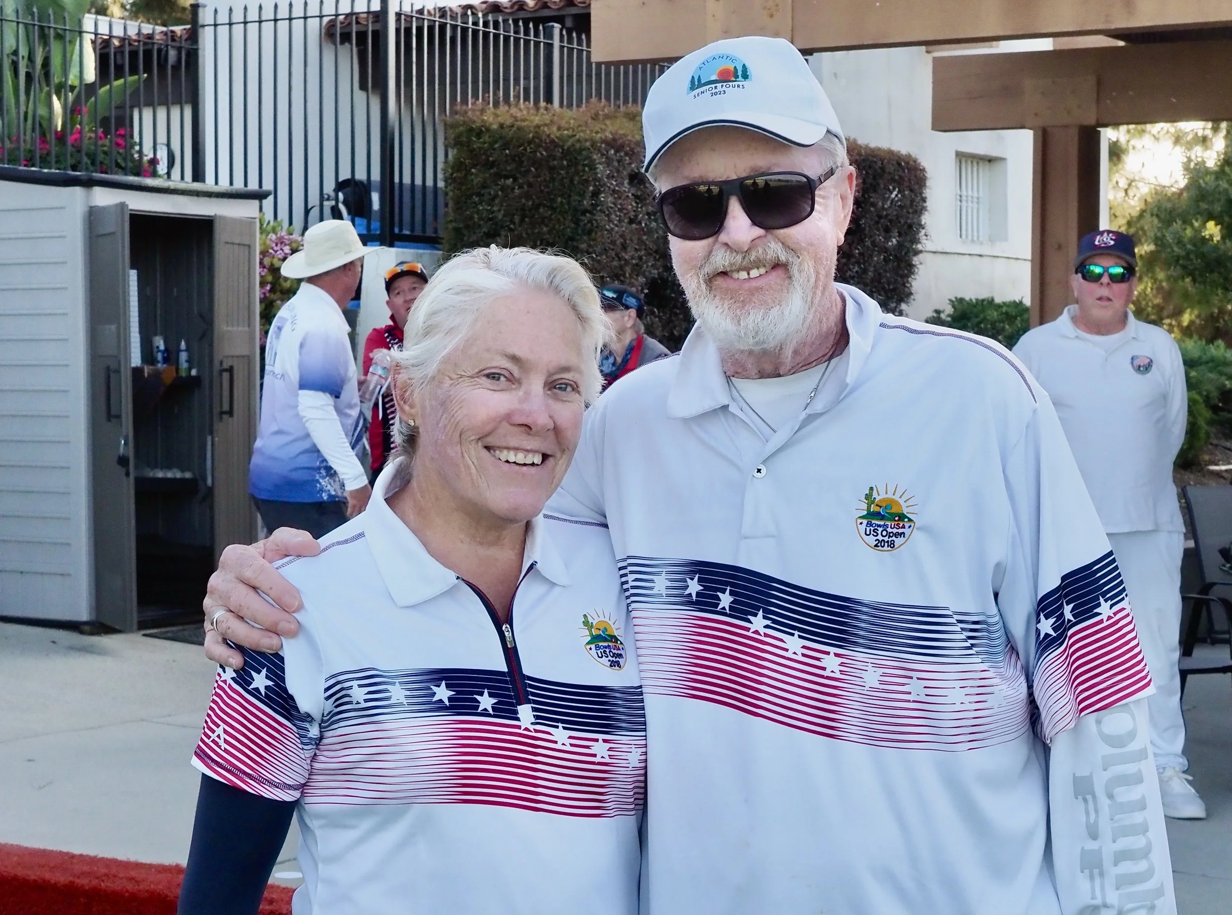 Two smiling people in white shirts with patriotic stripes and stars, one man and one woman, at a sports event. The man is wearing a cap, sunglasses, and a U.S. Open badge, while the woman has short blonde hair. In the background, several other people are gathered outdoors near a building with greenery.