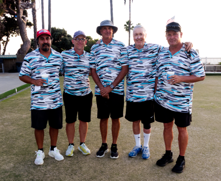 Five men in matching camouflage-pattern polo shirts and black shorts standing on a golf course, with palm trees and a clear sky in the background, holding drinks.