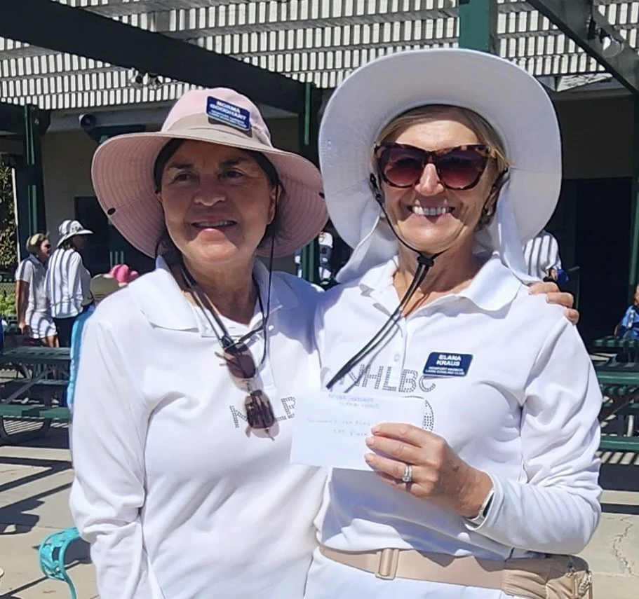 Two women smiling, wearing wide-brimmed hats and white shirts, at an outdoor event with people and benches in the background.