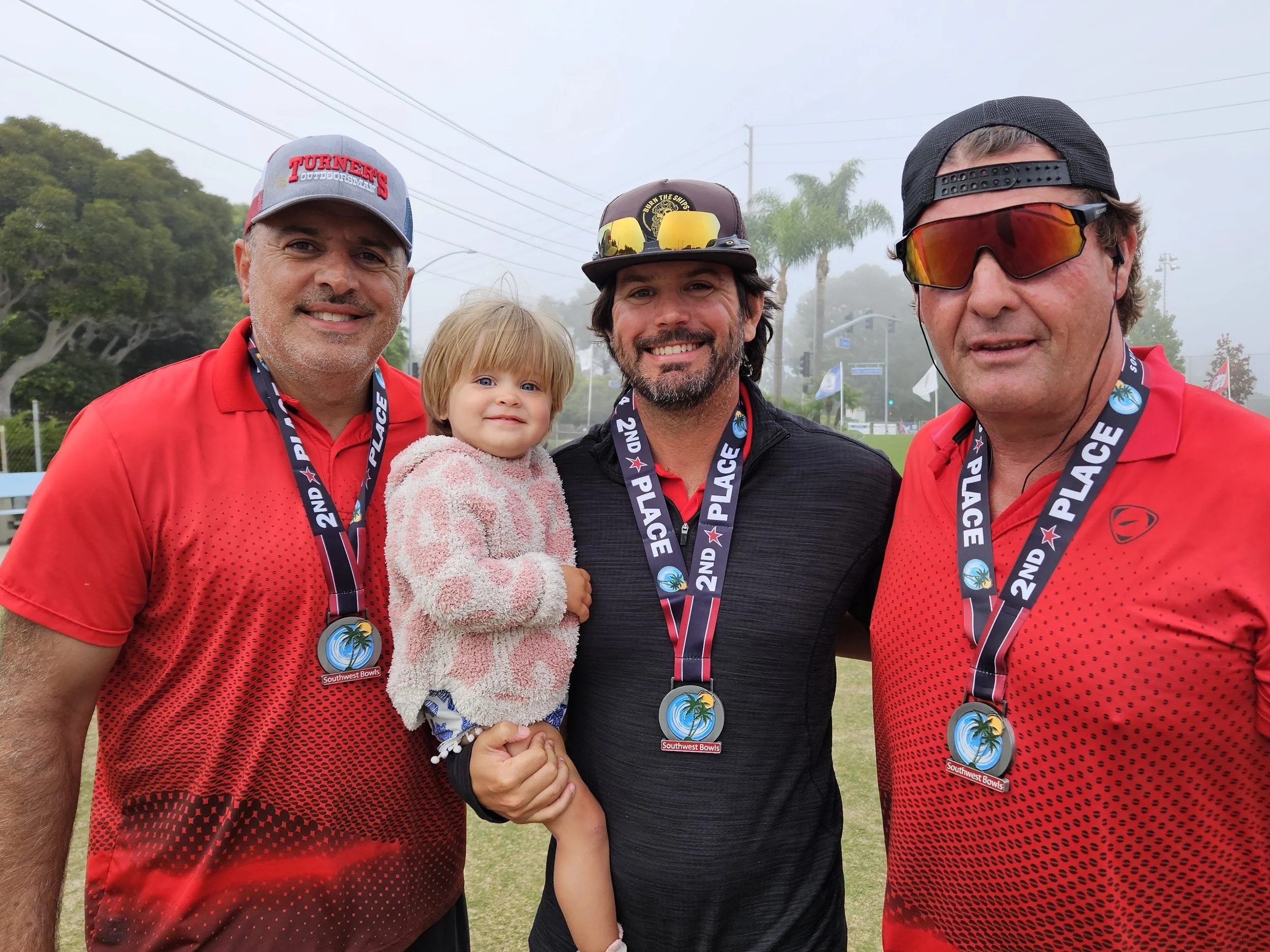 Three men and a young girl posing outdoors, all wearing medals that say '2nd Place' from the Southwest Bowls tournament, with a foggy background of trees and a sports field.