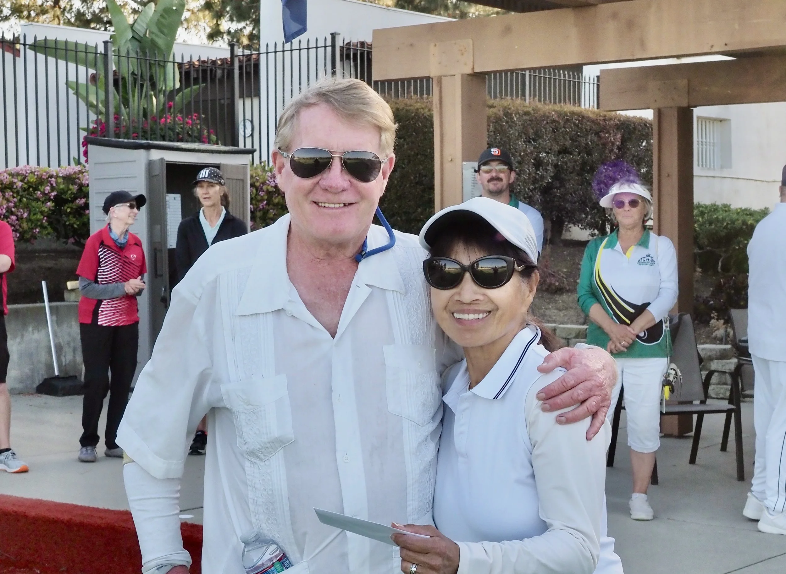 A man and a woman, both wearing sunglasses, are smiling and posing together at an outdoor event. The man is in a white shirt, and the woman is in a white visor and white shirt. In the background, several women are standing near a fence and flowering bushes, some wearing casual sportswear and hats.