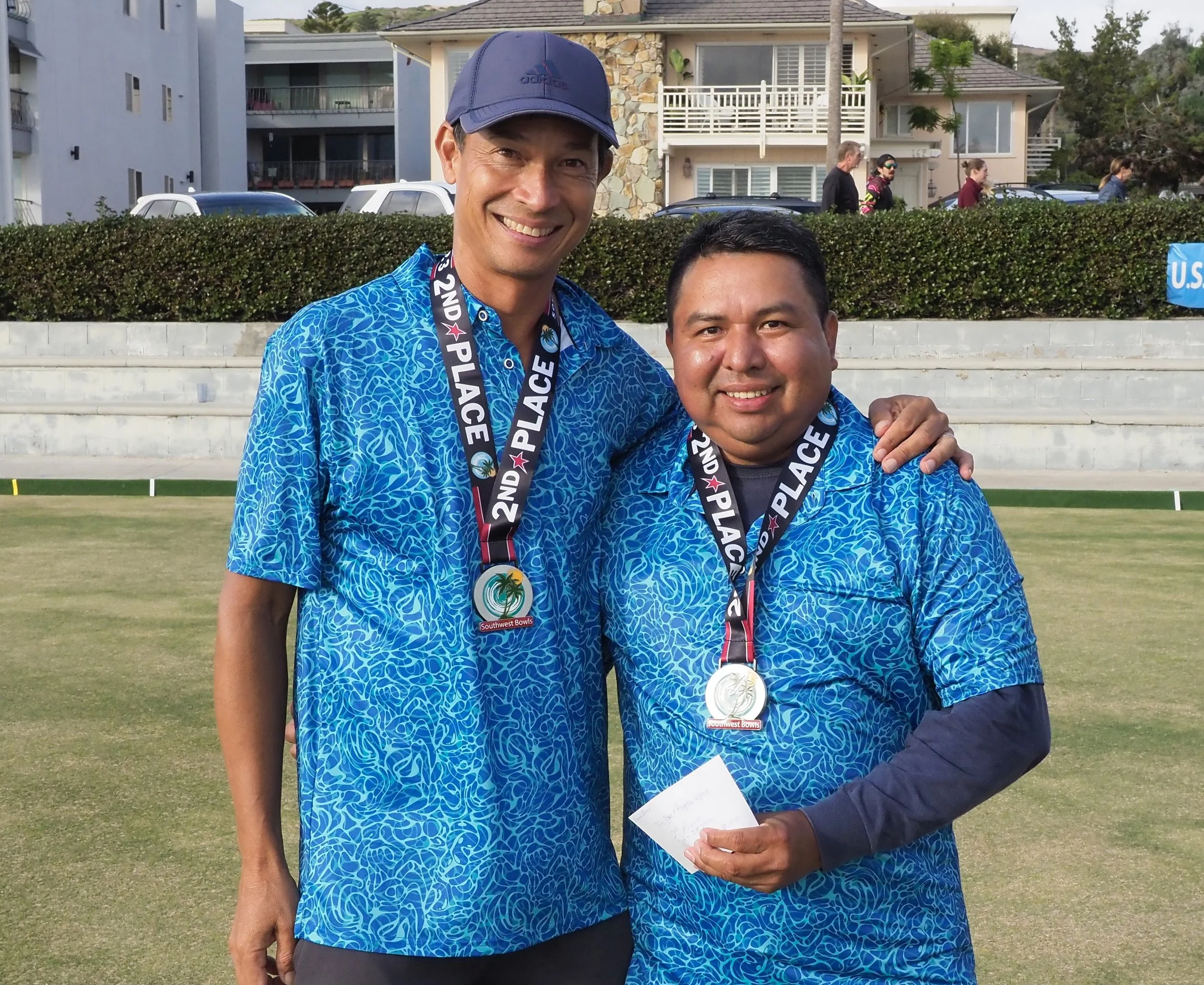 Two men wearing blue patterned shirts and medals around their necks standing on a grassy area, smiling for the camera. The medals indicate they placed second in a bowling competition, and a small paper is held by the man on the right.